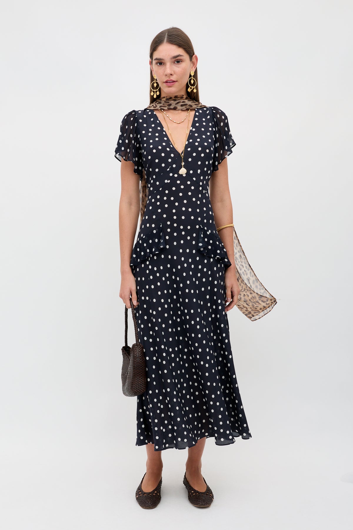 A woman in the Evie - Bohemia Spot Deep Navy dress with short sleeves, polka dots, a deep V-neck, styled with a sheer scarf, gold earrings, a brown handbag, and brown flats stands against a plain white background.