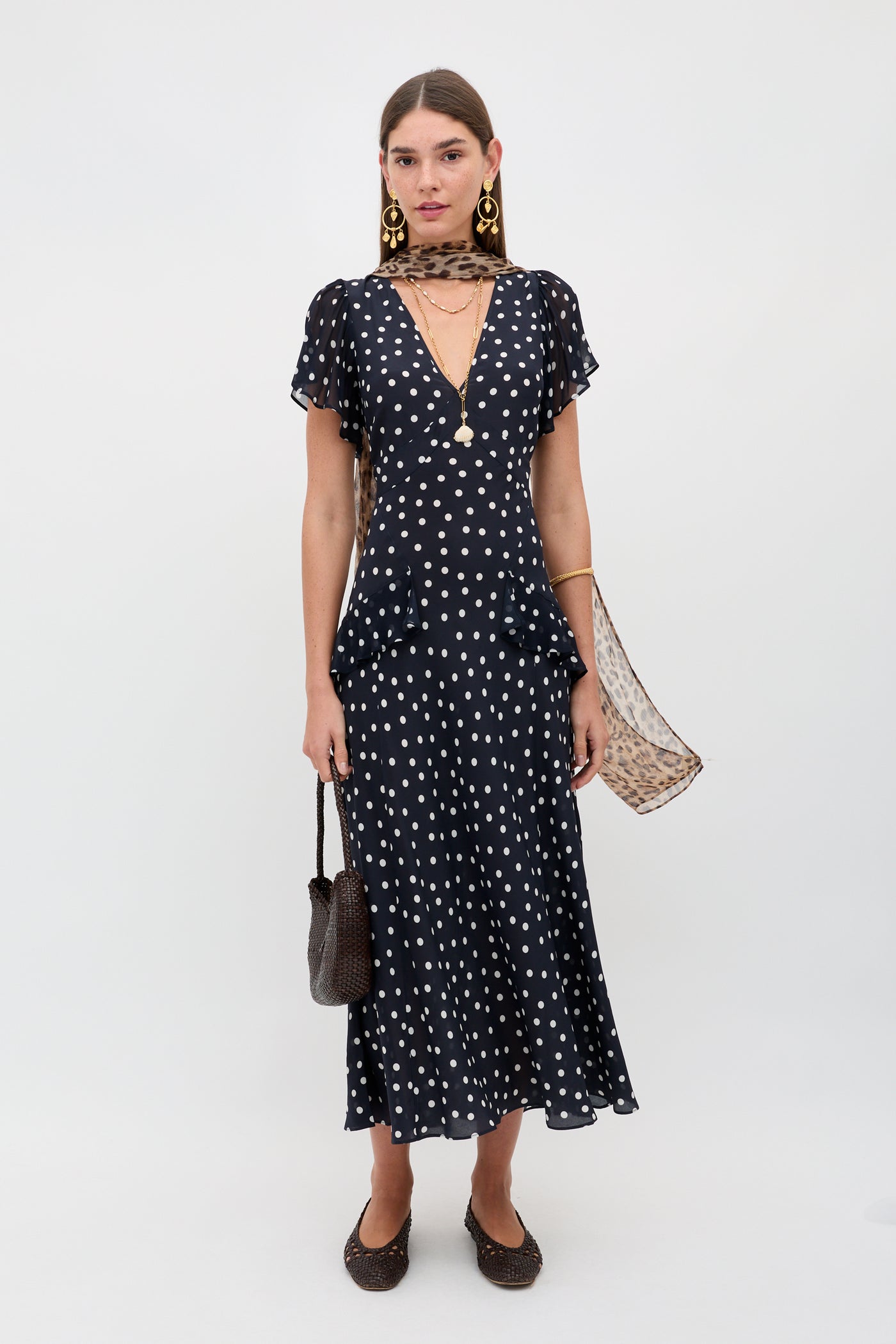 A woman in the Evie - Bohemia Spot Deep Navy dress with short sleeves, polka dots, a deep V-neck, styled with a sheer scarf, gold earrings, a brown handbag, and brown flats stands against a plain white background.