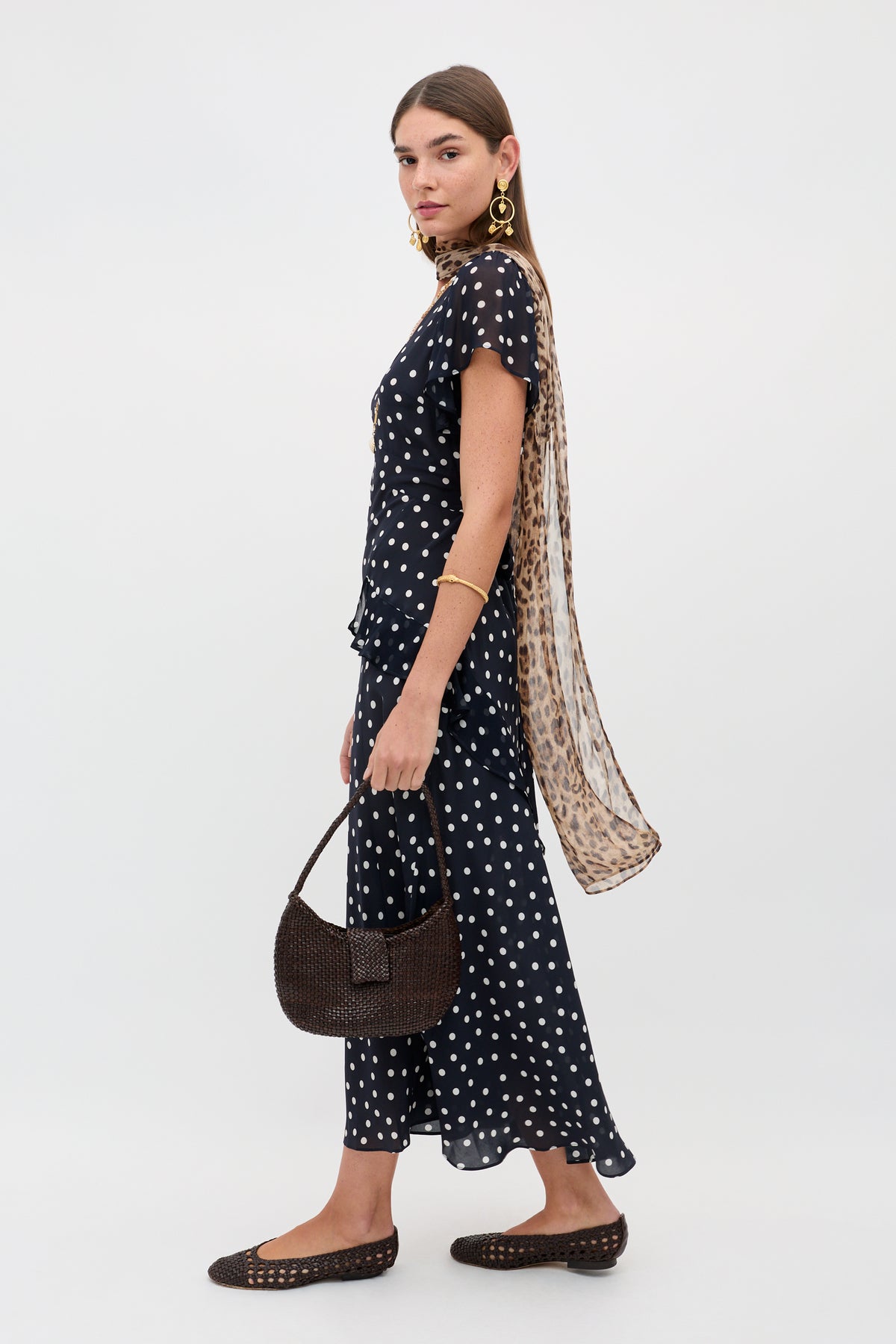 Woman in a black, short-sleeve polka dot top and matching skirt, brown flats, gold earrings, gold bracelet, leopard print scarf, and woven brown handbag—perfect for pairing with the Evie - Bohemia Spot Deep Navy dress for extra charm.