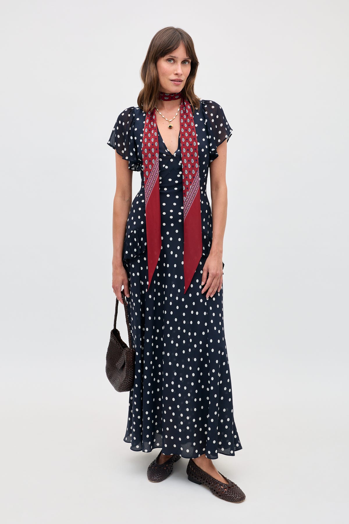A woman stands against a plain background wearing the Evie - Bohemia Spot Deep Navy dress. She pairs it with a maroon printed scarf, a woven brown bag, and matching woven brown flats.