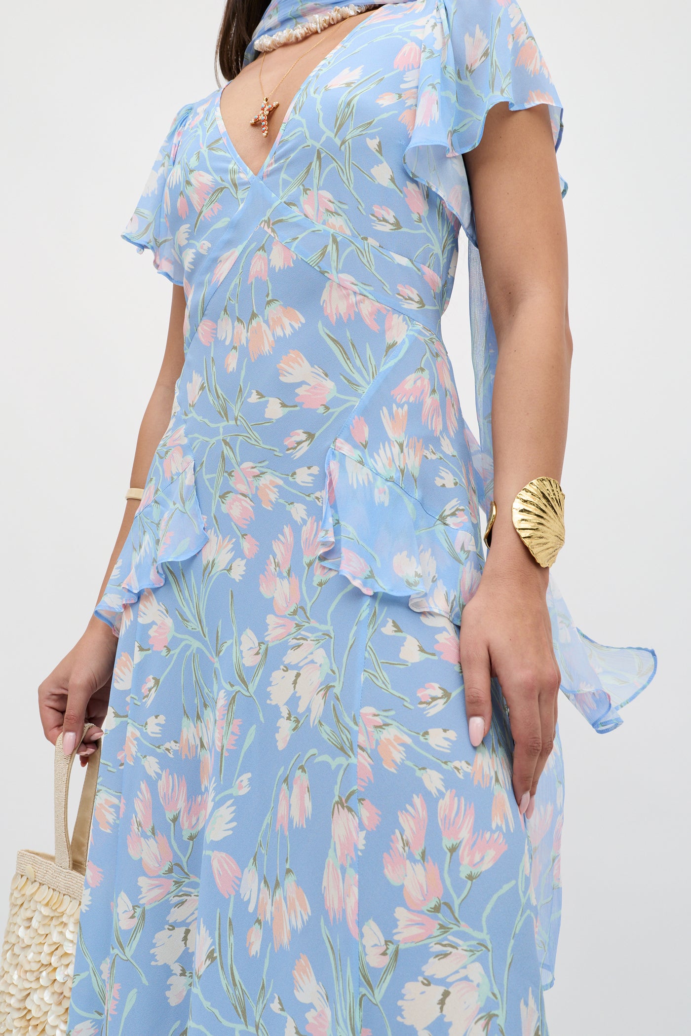 A woman wears the Evie - Watercolour Floral Blue dress with a gold shell-shaped bracelet, a necklace, and holds a woven straw bag. The image is cropped from the shoulders down.