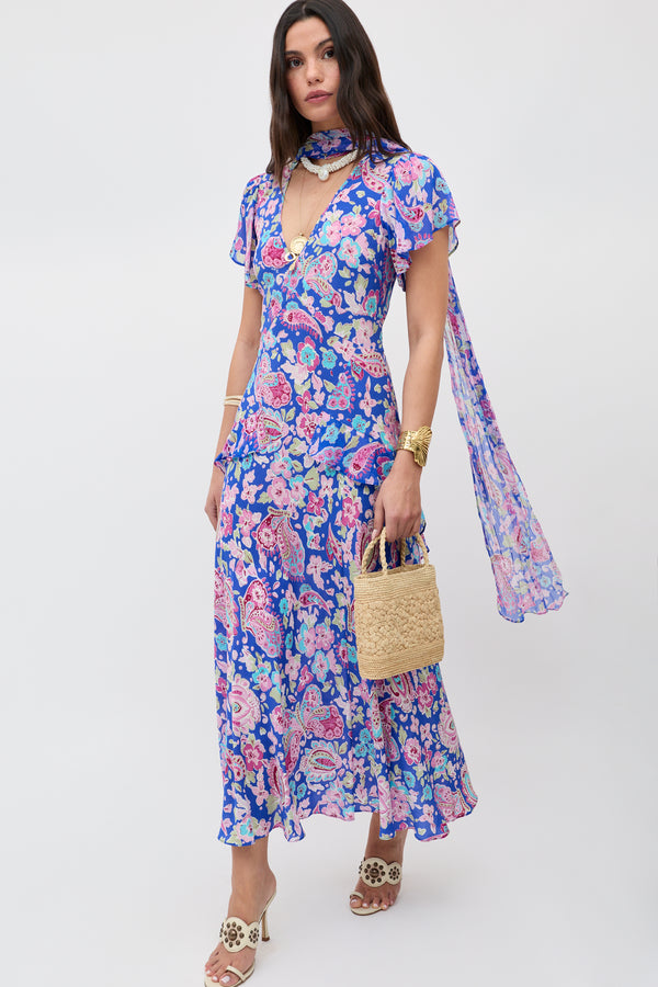 A woman in the Julien - Cara Floral Blue midi dress with short flutter sleeves and matching scarf, carrying a woven straw handbag and wearing white open-toe sandals with floral accents.