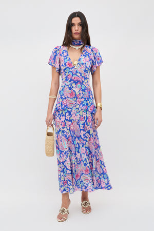 A woman with straight brown hair and a neutral expression wears the Evie - Cara Floral Blue maxi dress, beige sandals, gold jewelry, and holds a woven handbag against a white background.