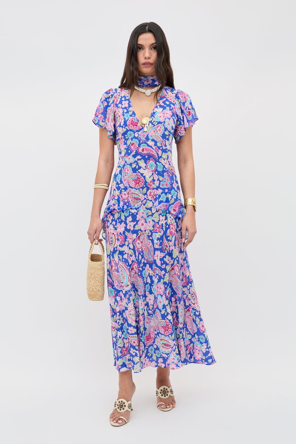 A woman with straight brown hair and a neutral expression wears the Evie - Cara Floral Blue maxi dress, beige sandals, gold jewelry, and holds a woven handbag against a white background.