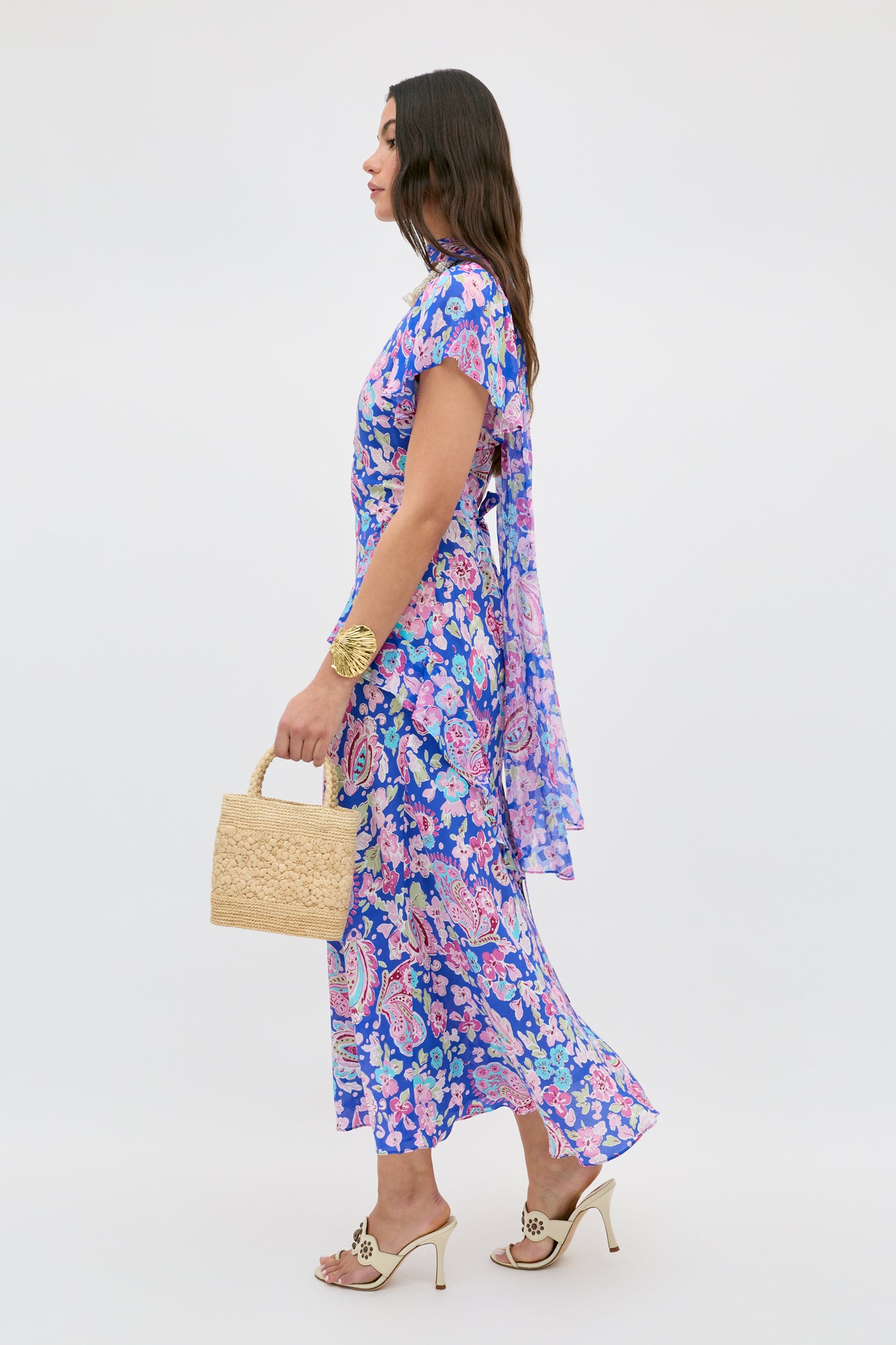 A woman models the Evie - Cara Floral Blue dress, featuring blue and purple florals and flutter sleeves. She poses in profile with a sheer scarf, straw handbag, high-heeled sandals, and long wavy hair against a plain white background.