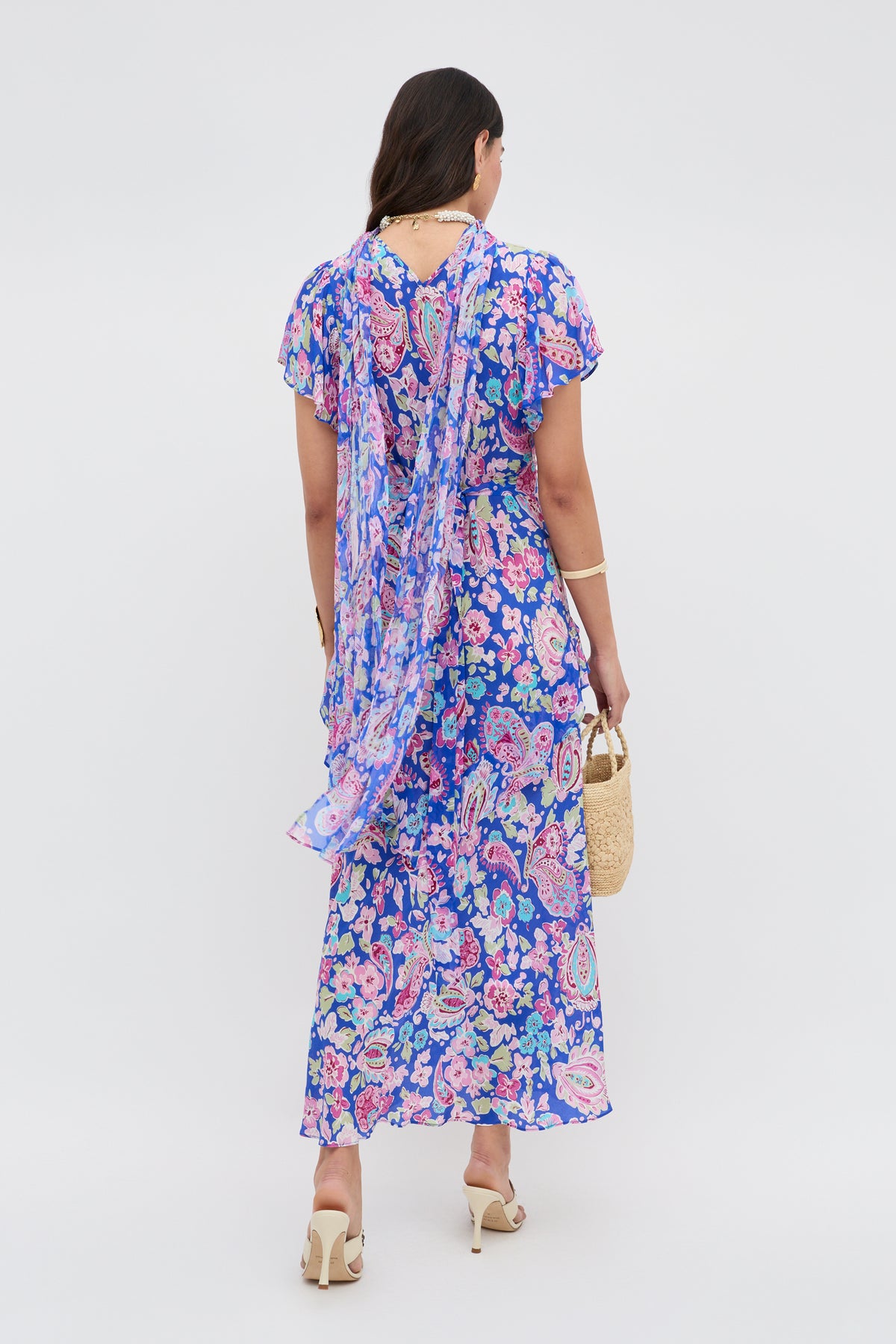 A woman faces away, wearing the Evie - Cara Floral Blue dress with sheer short sleeves, a long back scarf, and blue-pink floral patterns. She pairs it with a small woven handbag and cream high-heeled sandals.