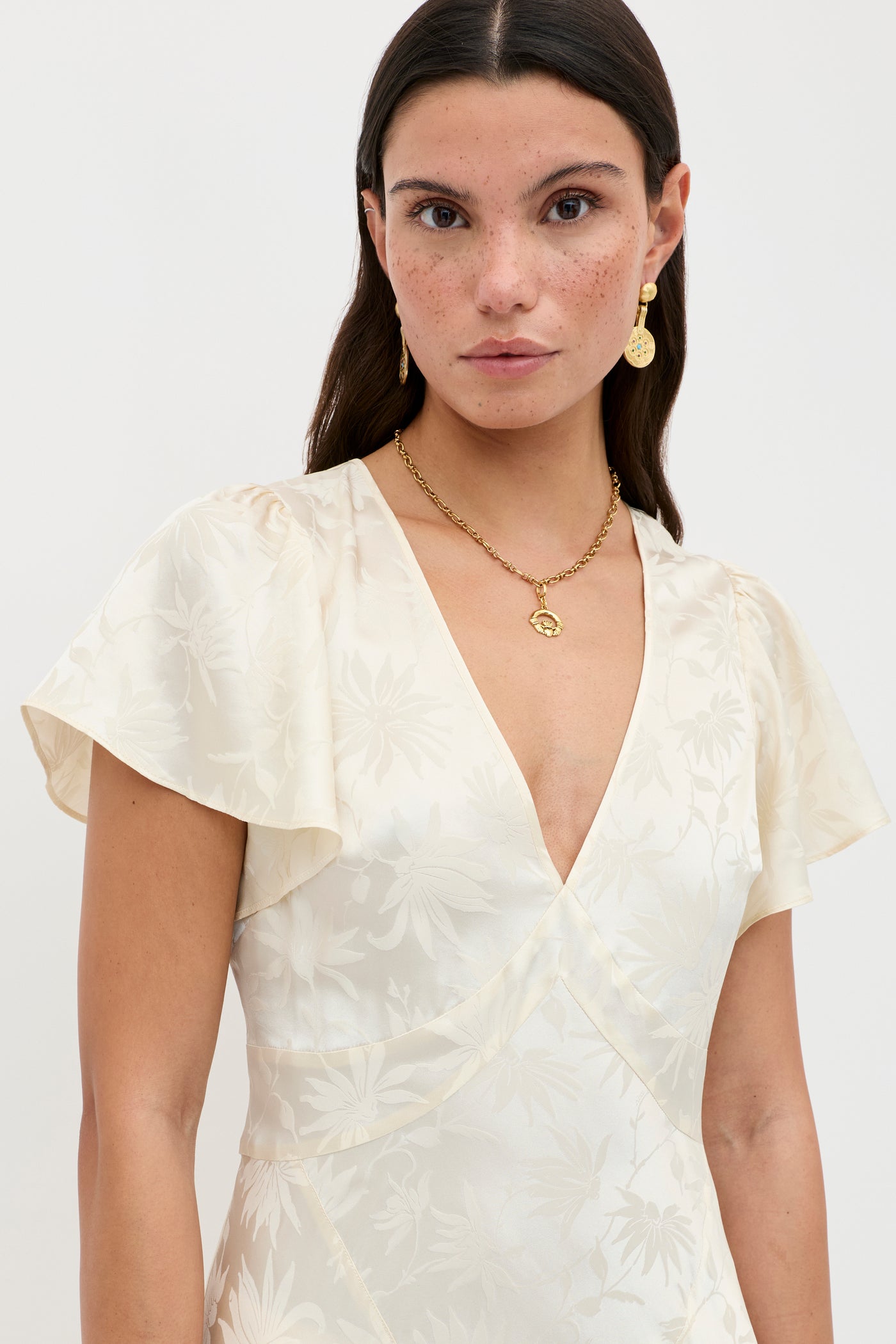 A woman with straight dark hair and freckles wears the Evie - Daisy Jacquard Champagne dress, featuring a cream floral pattern and short flutter sleeves, paired with gold earrings and a matching necklace against a plain light background.