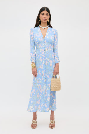 A woman in the "Katie - Watercolour Floral Blue" light blue, long-sleeved, floral midi dress with a v-neckline stands holding a woven handbag. She has long hair, wears sandals, gold jewelry, and a white beaded necklace.