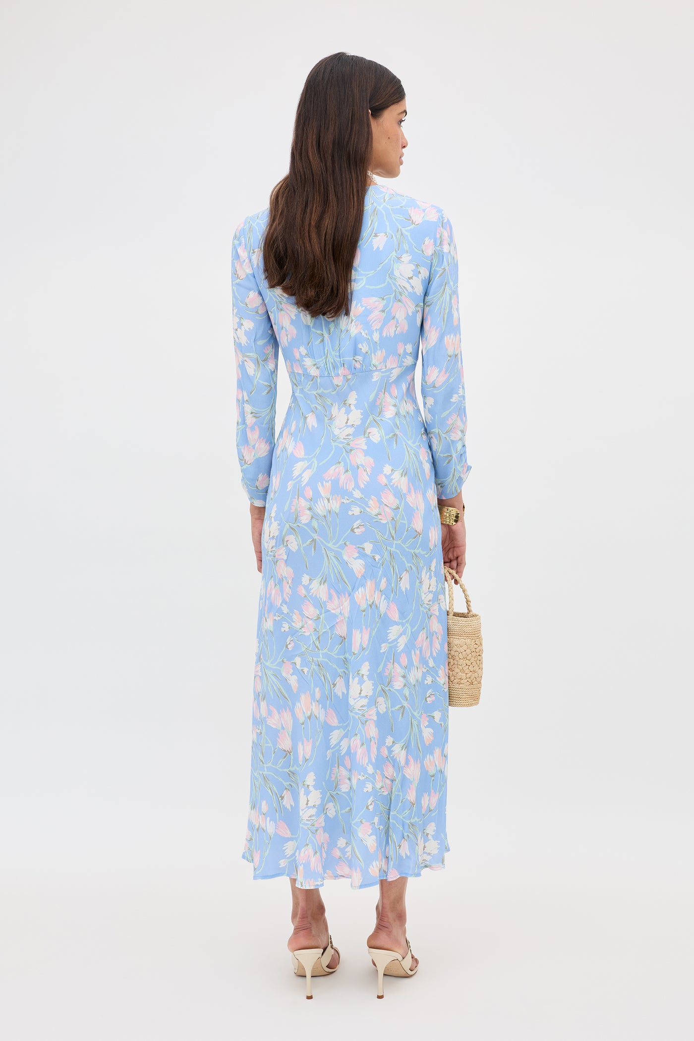 A woman with long brown hair faces away, wearing the Katie - Watercolour Floral Blue midi dress with long sleeves and beige heels. She holds a small woven bag, standing against a plain white background.
