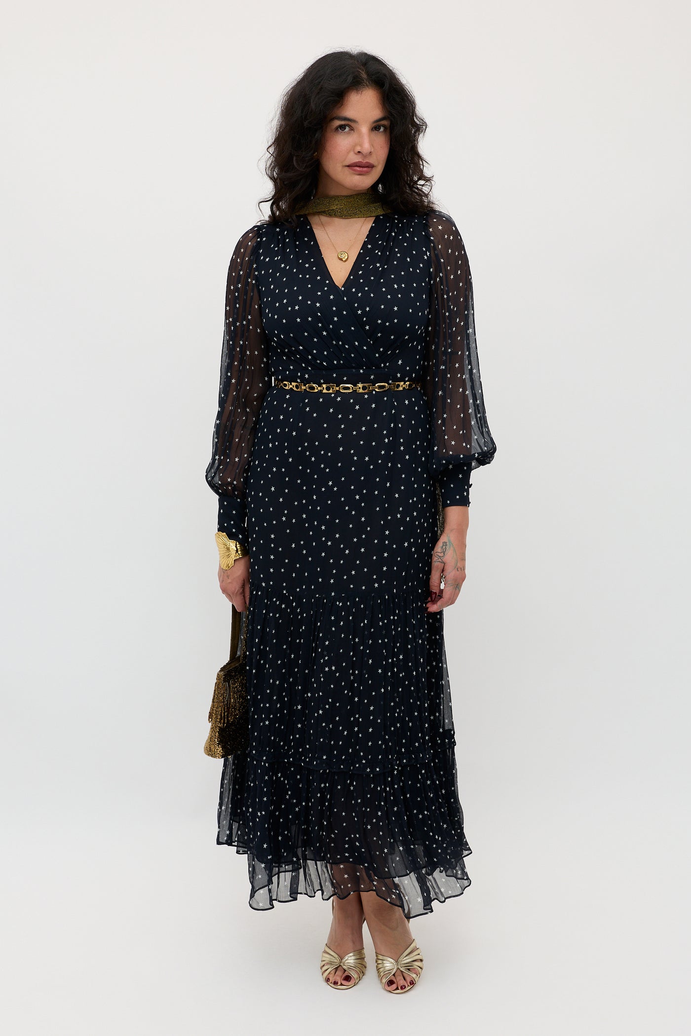 A woman stands against a plain background in the Elsie - Starry Night Navy dress—a long, sheer-sleeved navy dress with white dots—paired with gold accessories and open-toed shoes. Her wavy dark hair frames her neutral expression.