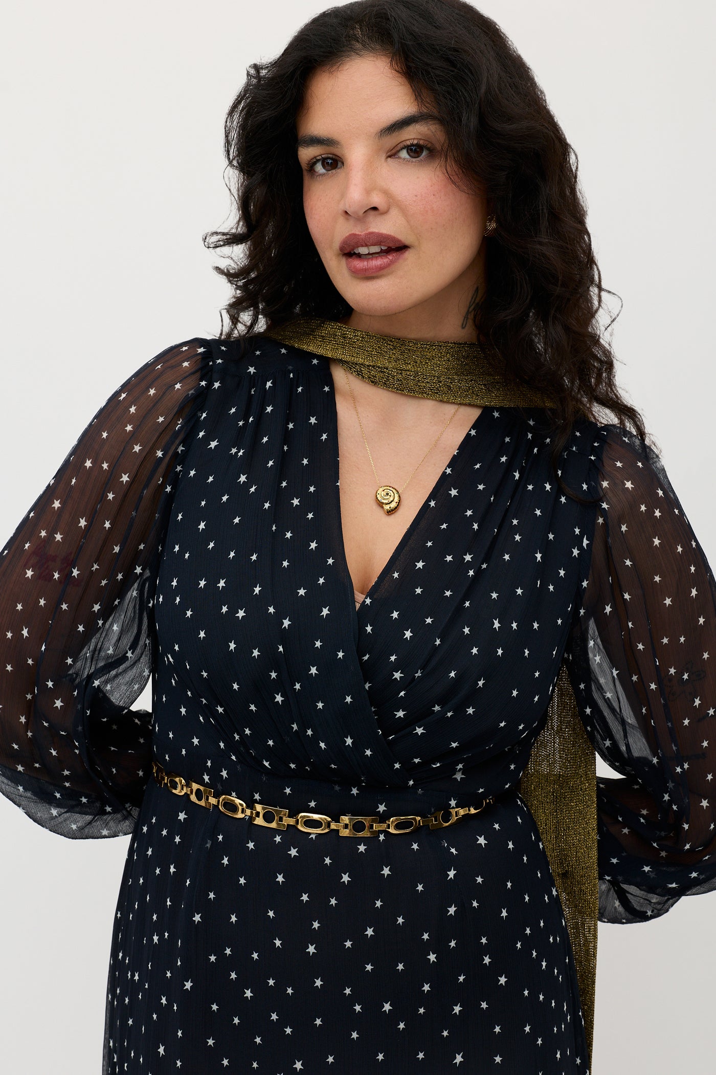 A woman with long, wavy dark hair wears the Elsie - Starry Night Navy dress—navy with white stars, sheer puffed sleeves, a gold chain belt and necklace, plus a sparkly gold scarf—standing against a plain background.