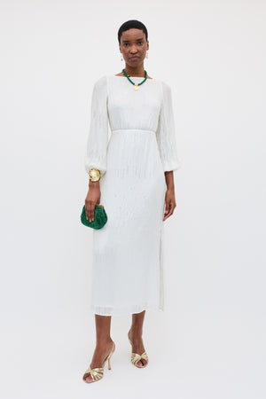 A woman in the Coco - Ivory vintage wedding gown with a bias-cut skirt and sheer sleeves, gold heels, a chunky green necklace, gold bracelet, and a small green clutch stands against a plain white background.