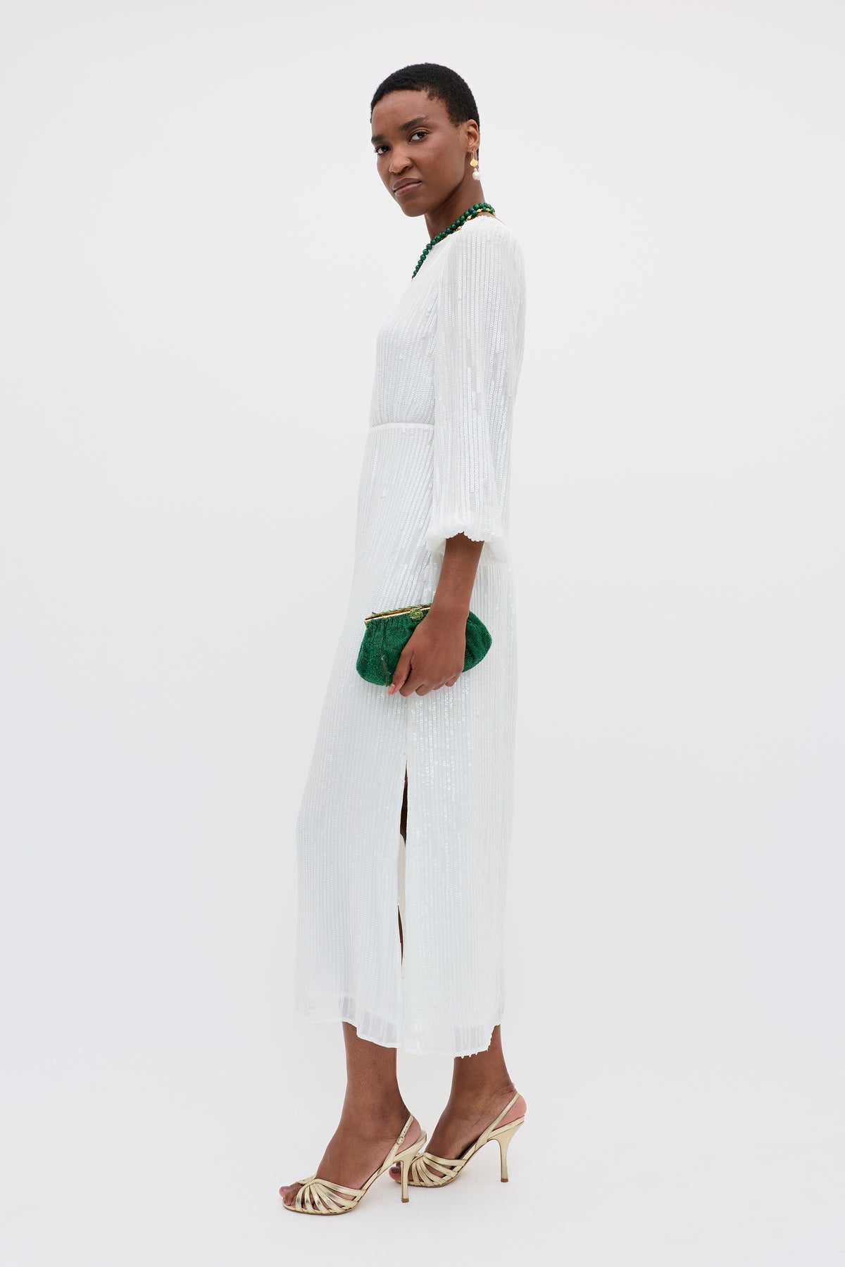 Wearing the Coco - Ivory sequin dress with a bias-cut skirt, gold heels, a green beaded necklace, and holding a green clutch, a person stands against a plain white background.