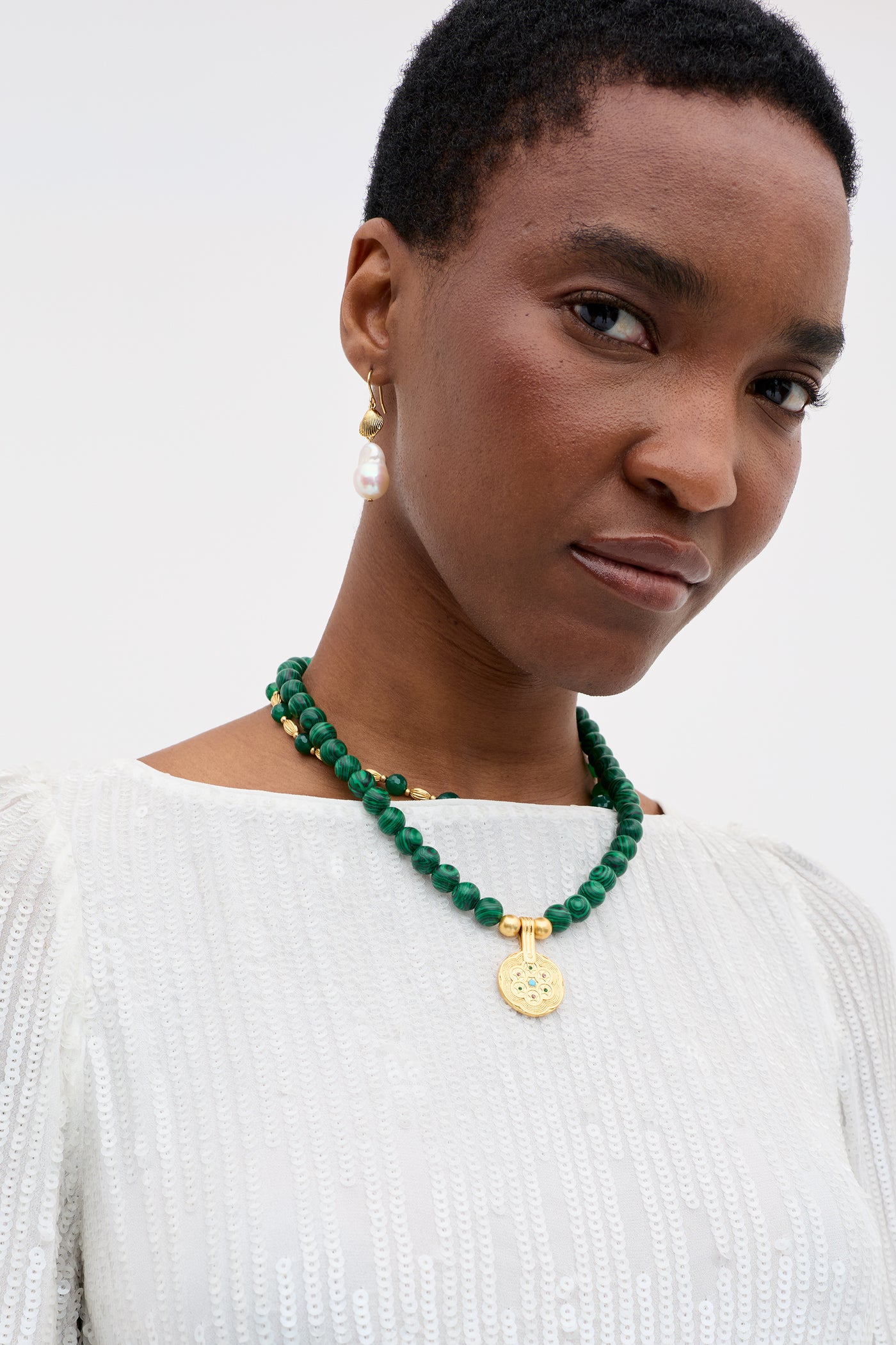 A woman with short hair wears the Coco - Ivory white textured top, a green beaded necklace with gold pendant, and a single dangling pearl earring, gazing neutrally into the camera.