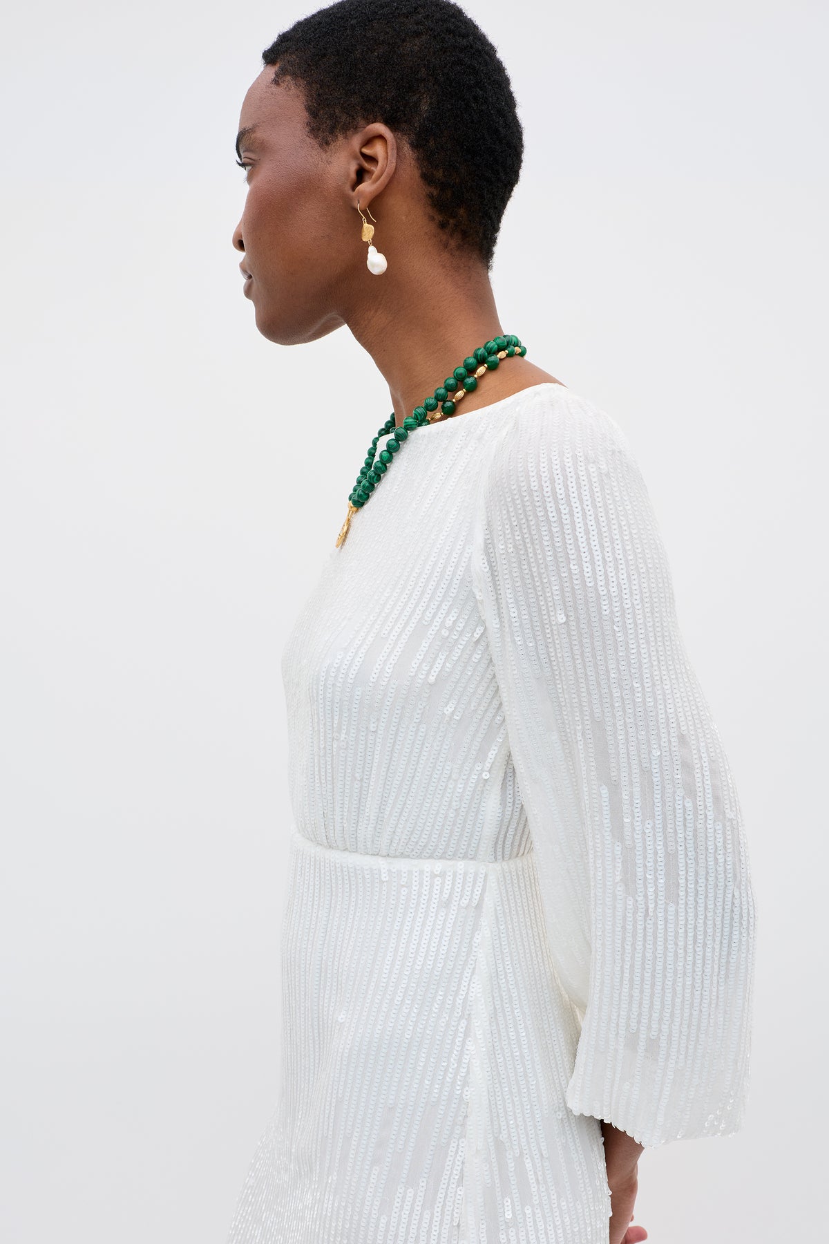 A person with short hair wears the Coco - Ivory long-sleeved, white, textured vintage wedding gown with gold earrings and a green beaded necklace, shown in profile against a plain light background.