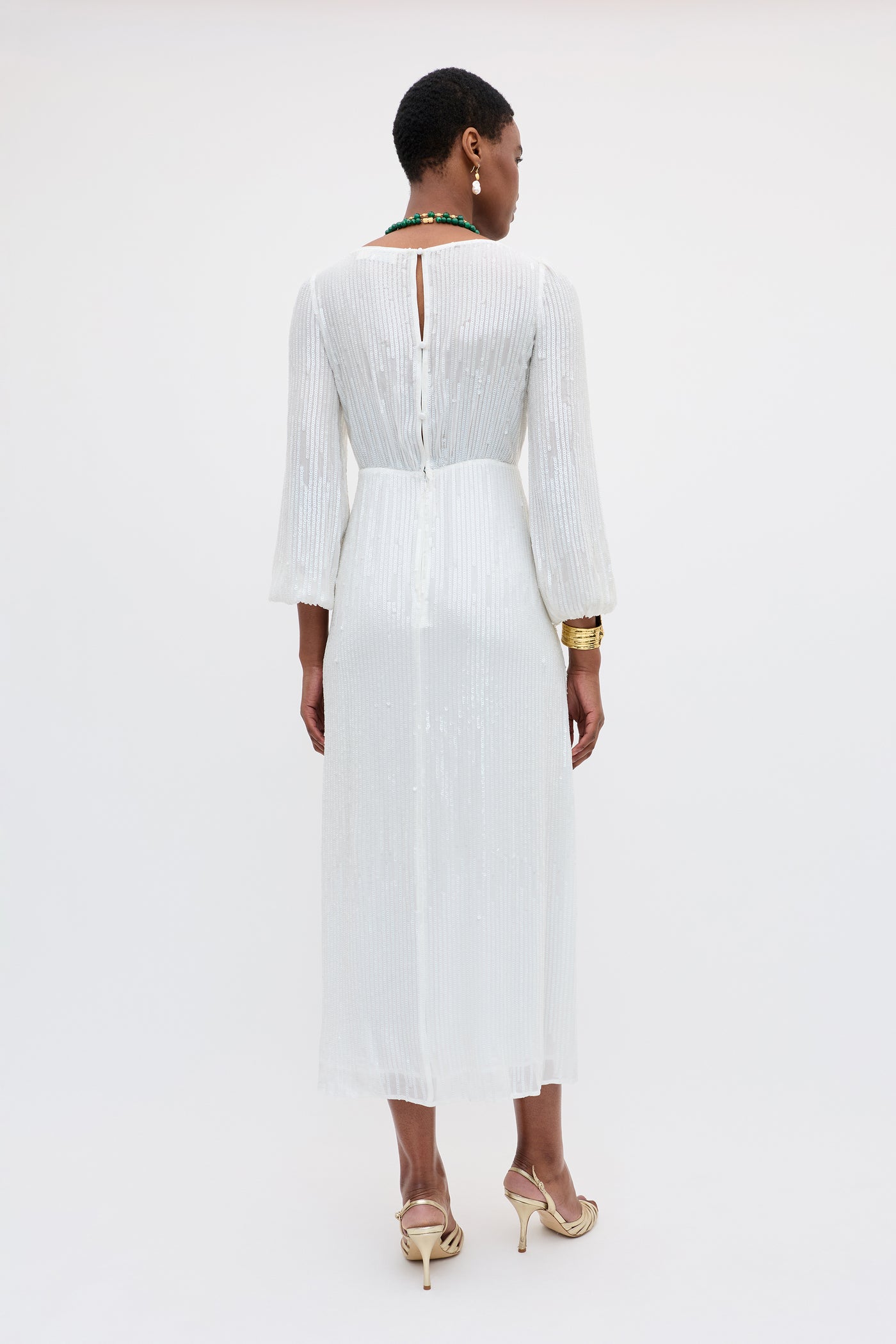 A woman facing away wears the Coco - Ivory dress, an ivory sequin gown with vertical pleats, a keyhole back, and long sleeves. She pairs it with gold sandals, a green necklace, and gold bracelets against a plain white background.