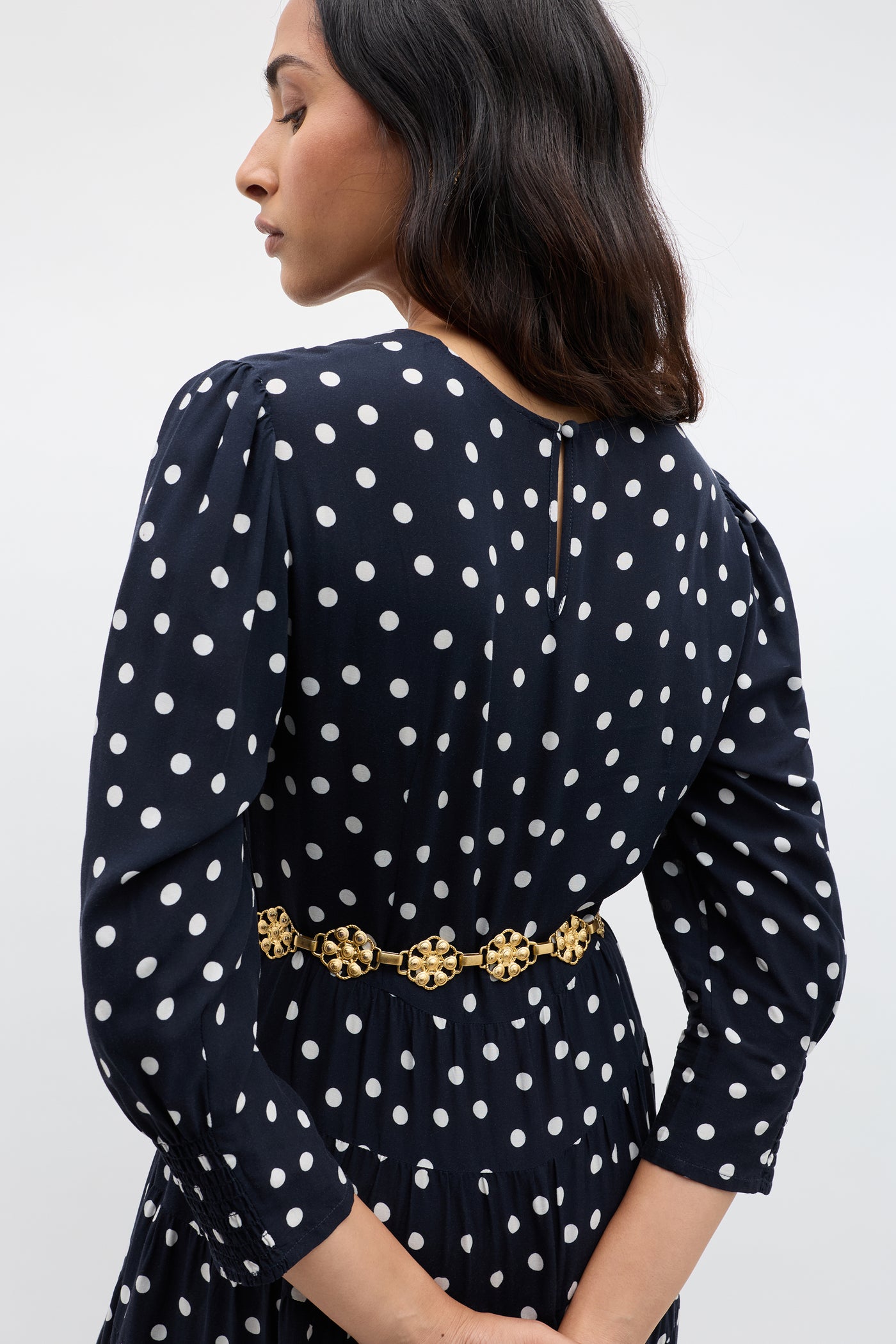A woman with long dark hair is seen from the back, wearing the Kristen - Bohemia Spot Navy dress, featuring white spots and a gold decorative belt. She turns her head slightly against a plain, light-colored background.