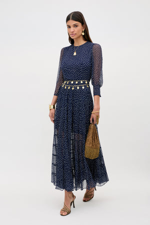 Woman wearing the Kristen - Navy Polka Dot georgette tiered-skirt dress, styled with a gold belt, jewelry, heeled sandals, and a woven brown purse, standing against a plain light background.