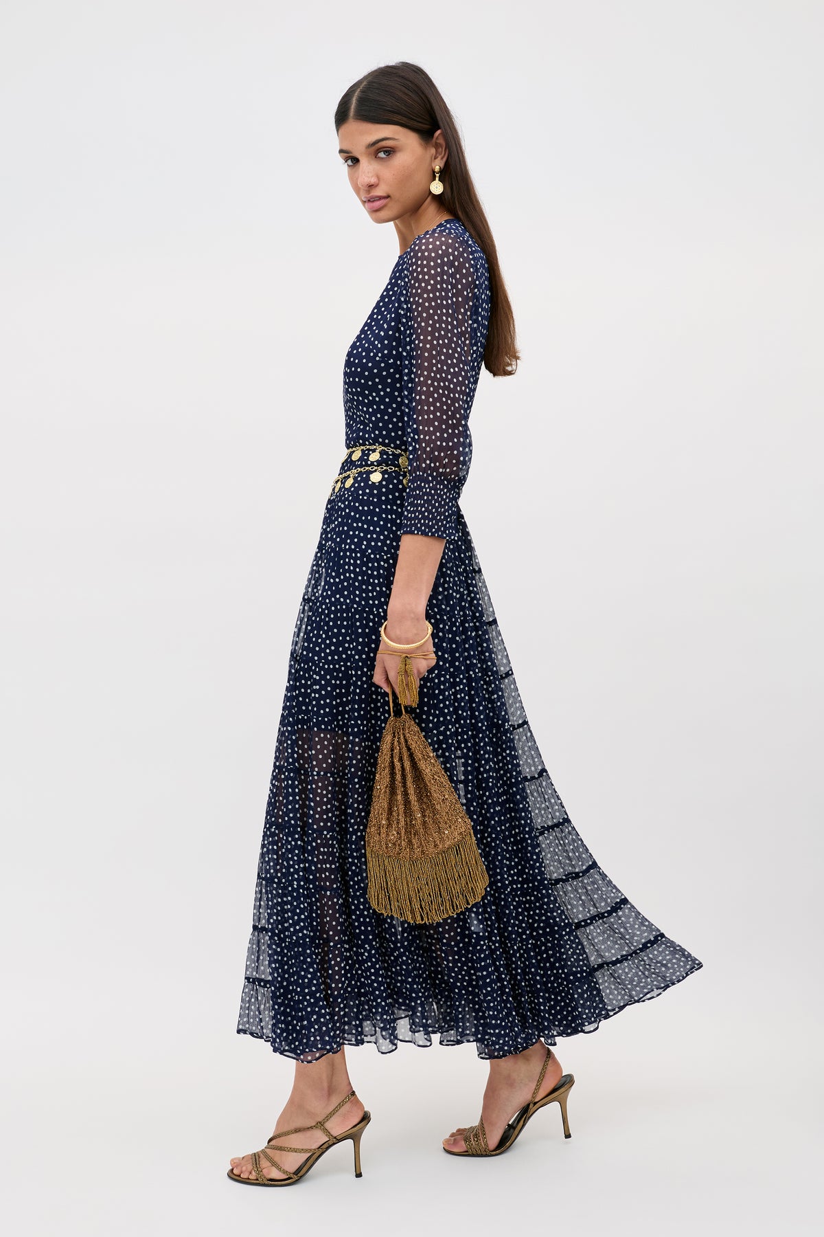 A woman in the Kristen - Navy Polka Dot long-sleeved tiered georgette dress with gold heels, a gold textured handbag, and gold jewelry stands against a plain white background.