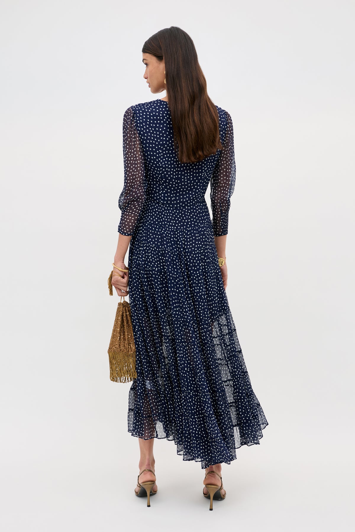A woman faces away, wearing the Kristen - Navy Polka Dot ankle-length georgette dress with an ivory spot print. She carries a textured brown handbag, wears heeled sandals, and has long brown hair down. The background is plain.