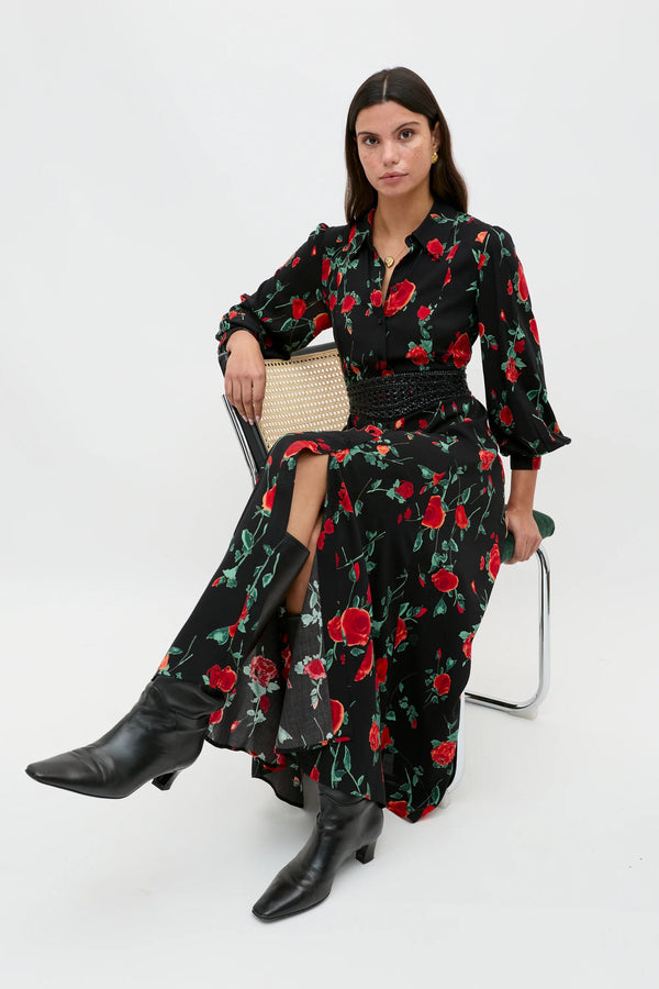 A petite woman with straight dark hair and a neutral expression wears the Maddison - Claudia Rose Black long shirt dress, featuring a red rose pattern and black belt, paired with heeled boots, set against a plain white background.