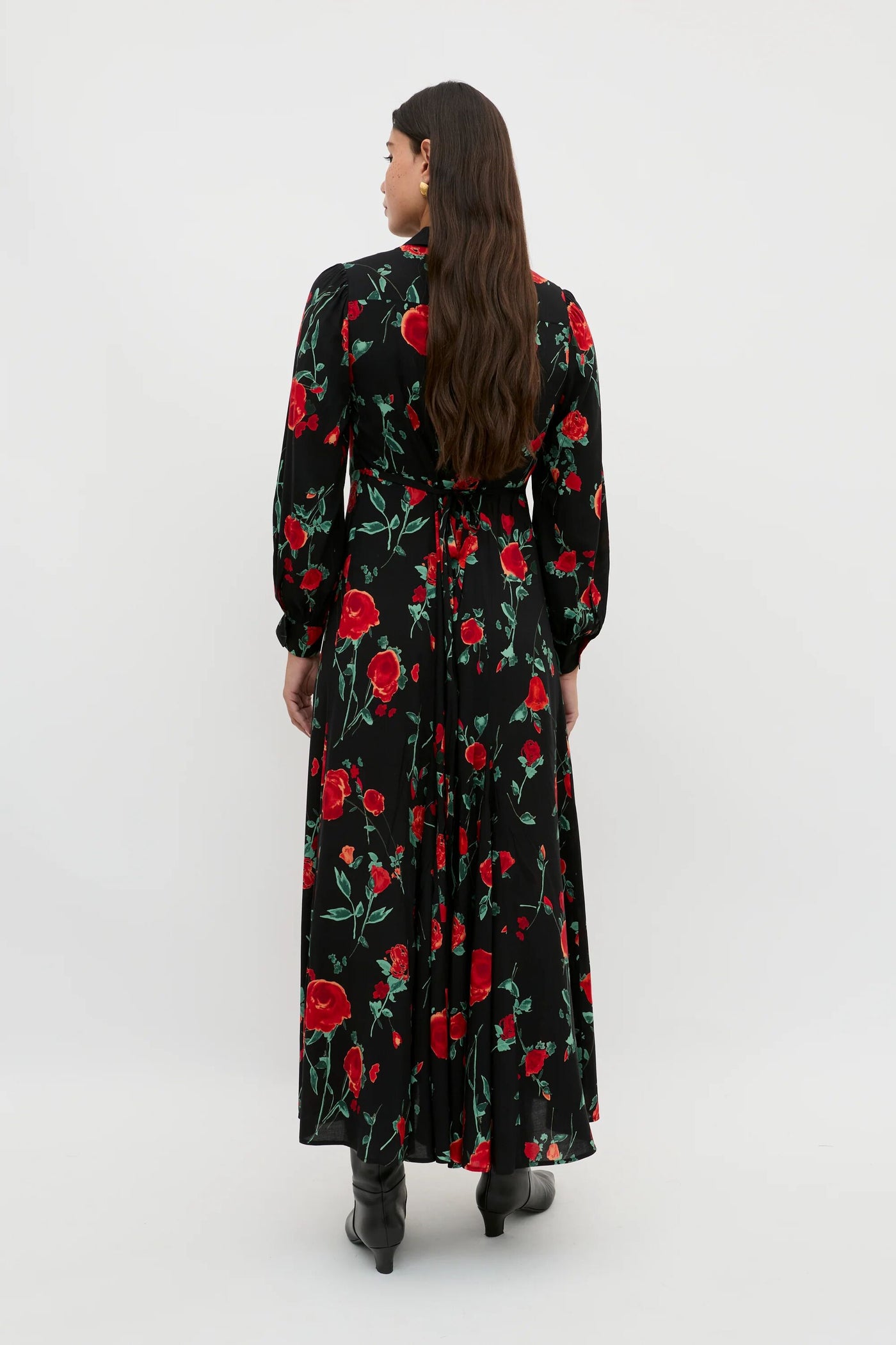 A petite person with long brown hair stands facing away against a plain light background, wearing the Maddison - Claudia Rose Black shirt dress with a red floral pattern and black boots.