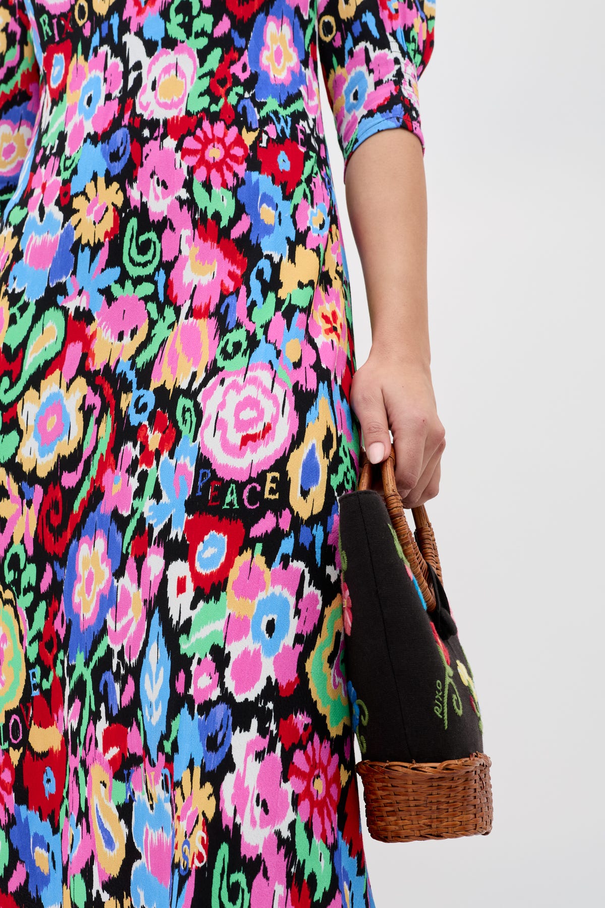 A person in the Jess - Ikat Bloom Charcoal dress, featuring vibrant pink, blue, yellow, and green florals on black with PEACE and LOVE prints, holds a small wicker basket bag.