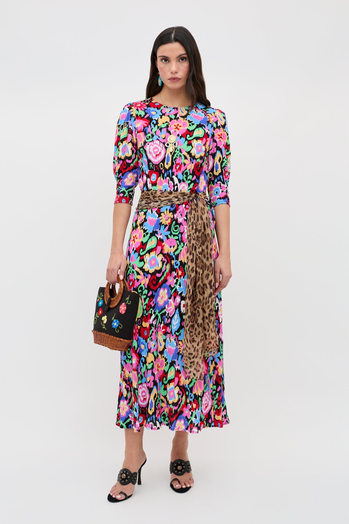 A woman stands against a plain background wearing the Jess - Ikat Bloom Charcoal dress, featuring puffed sleeves and a leopard print sash, holding a black floral-detailed handbag and wearing black heeled sandals.