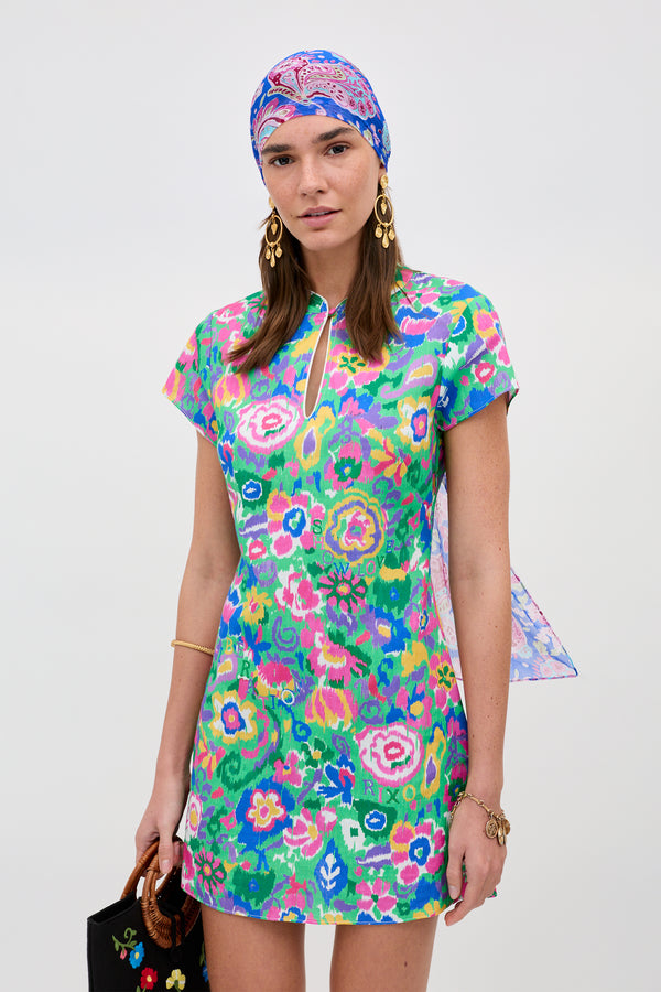 A woman poses against a plain light background wearing the Lolita - Ikat Bloom Green mini dress and matching headscarf, accessorized with gold earrings, bracelets, and a black handbag with floral details.
