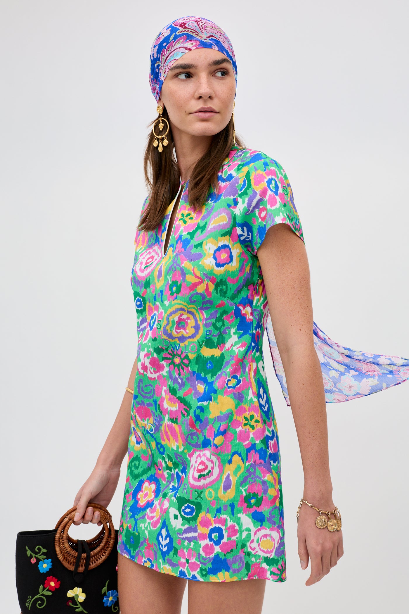 A woman in the Lolita - Ikat Bloom Green floral dress with a matching headscarf holds an embroidered black handbag. She wears gold jewelry and looks to the side against a plain background.