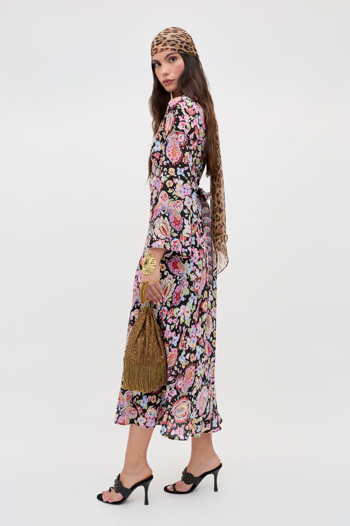 A woman models the Tania - Cara Floral Black dress, paired with black heeled sandals and a leopard-print headscarf, holding a gold beaded handbag against a plain white background.
