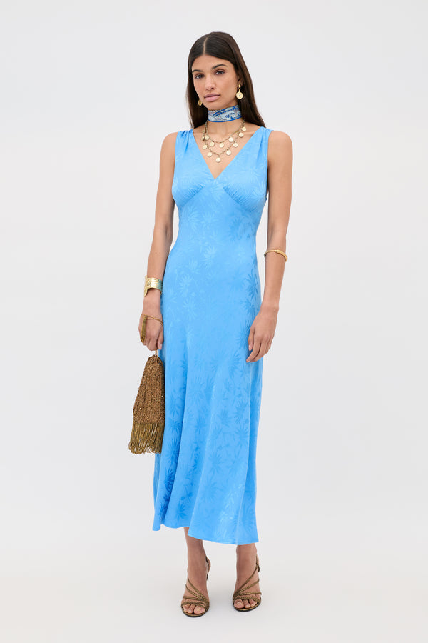 A woman in the Sandrine - Daisy Jacquard Blue V-neck midi slip dress with gold jewelry, a gold scarf, strappy heels, and a fringed gold handbag stands against a plain white background.