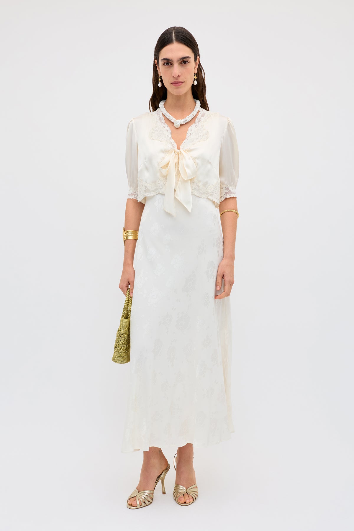 A woman in the Sandrine - Poppy, a vintage thirties ivory jacquard dress with lace and a large bow neckline, gold sandals, jewelry, and a small gold bag stands against a plain white background.
