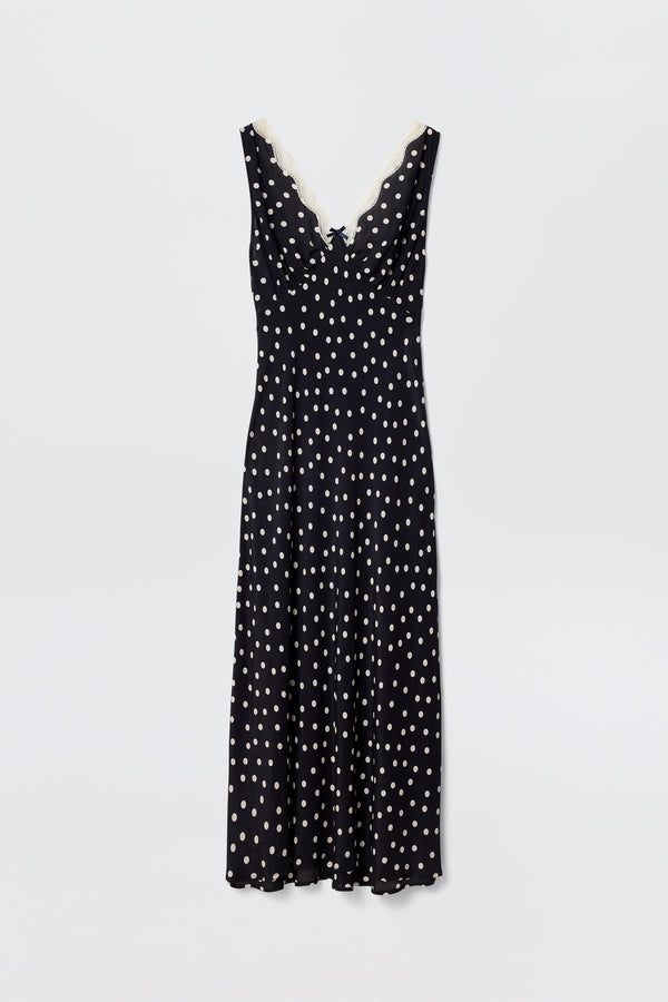 The Sandrine – Bohemia Spot Deep Navy is a sleeveless slip dress in deep navy with white polka dots, a V-neckline trimmed with white lace, and a flattering bias-cut midi hem, shown against a plain white background.