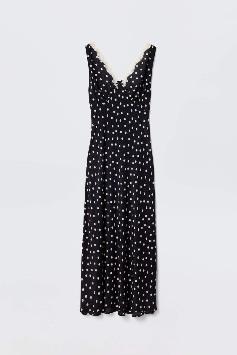 The Sandrine – Bohemia Spot Deep Navy is a sleeveless slip dress in deep navy with white polka dots, a V-neckline trimmed with white lace, and a flattering bias-cut midi hem, shown against a plain white background.