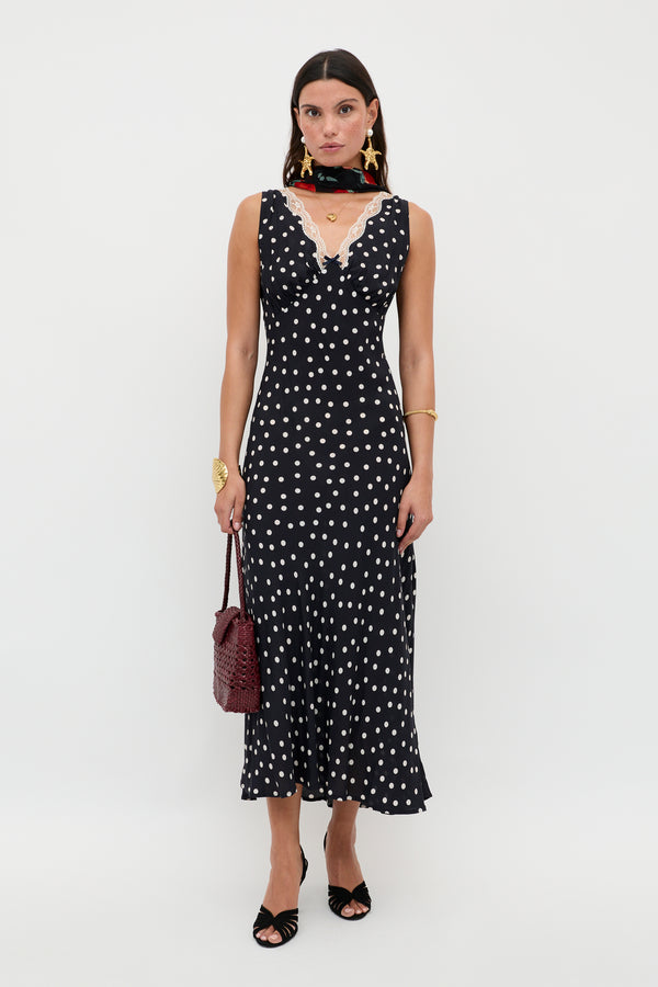 A woman stands against a white background wearing the Sandrine - Bohemia Spot Deep Navy dress with black open-toe heels, gold jewelry, a black scarf, and holding a textured brown handbag.