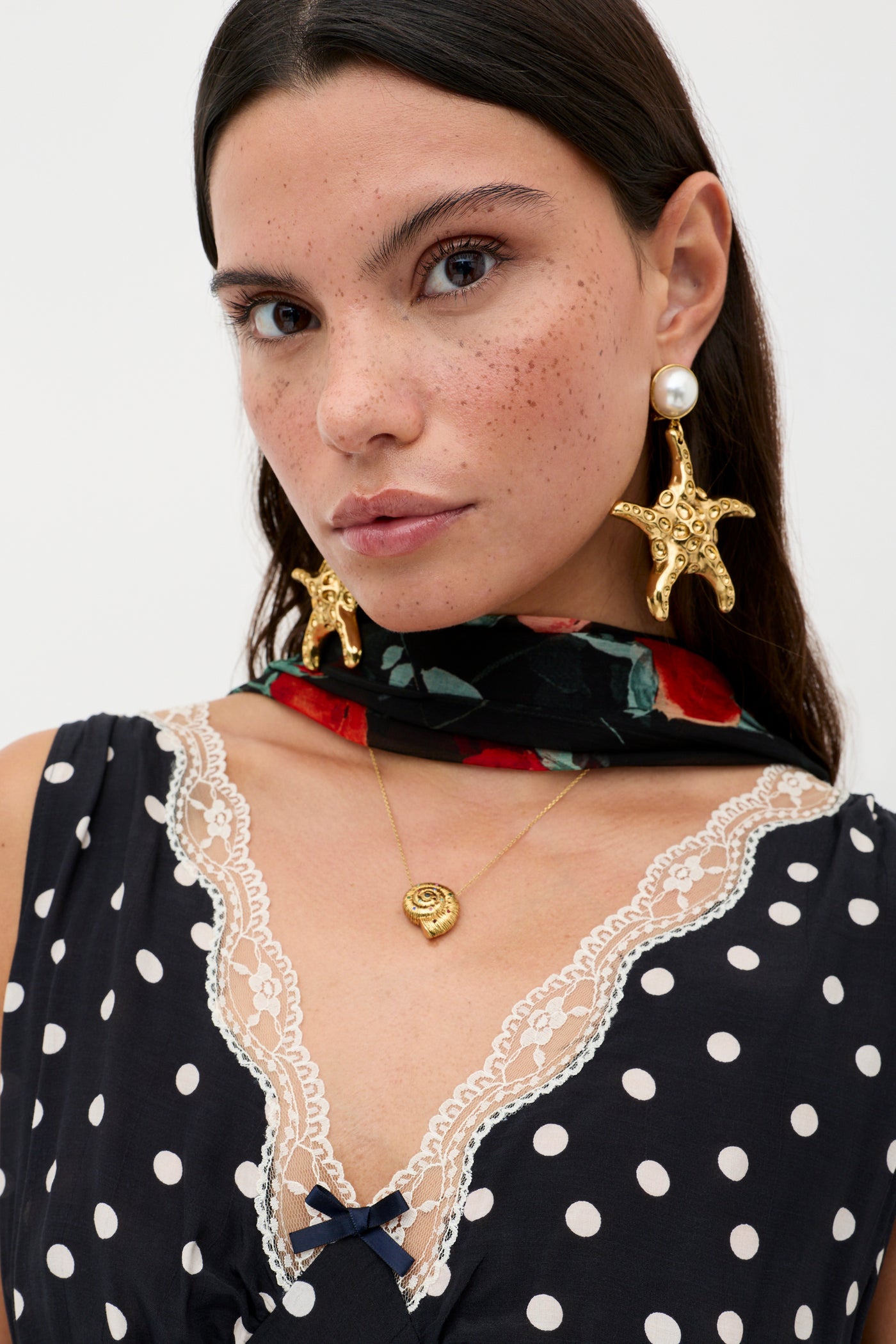 A woman with long brown hair wears gold starfish earrings, a gold pendant necklace, a floral scarf, and the RIXO Sandrine - Bohemia Spot Deep Navy slip dress with lace trim as she looks at the camera against a plain background.