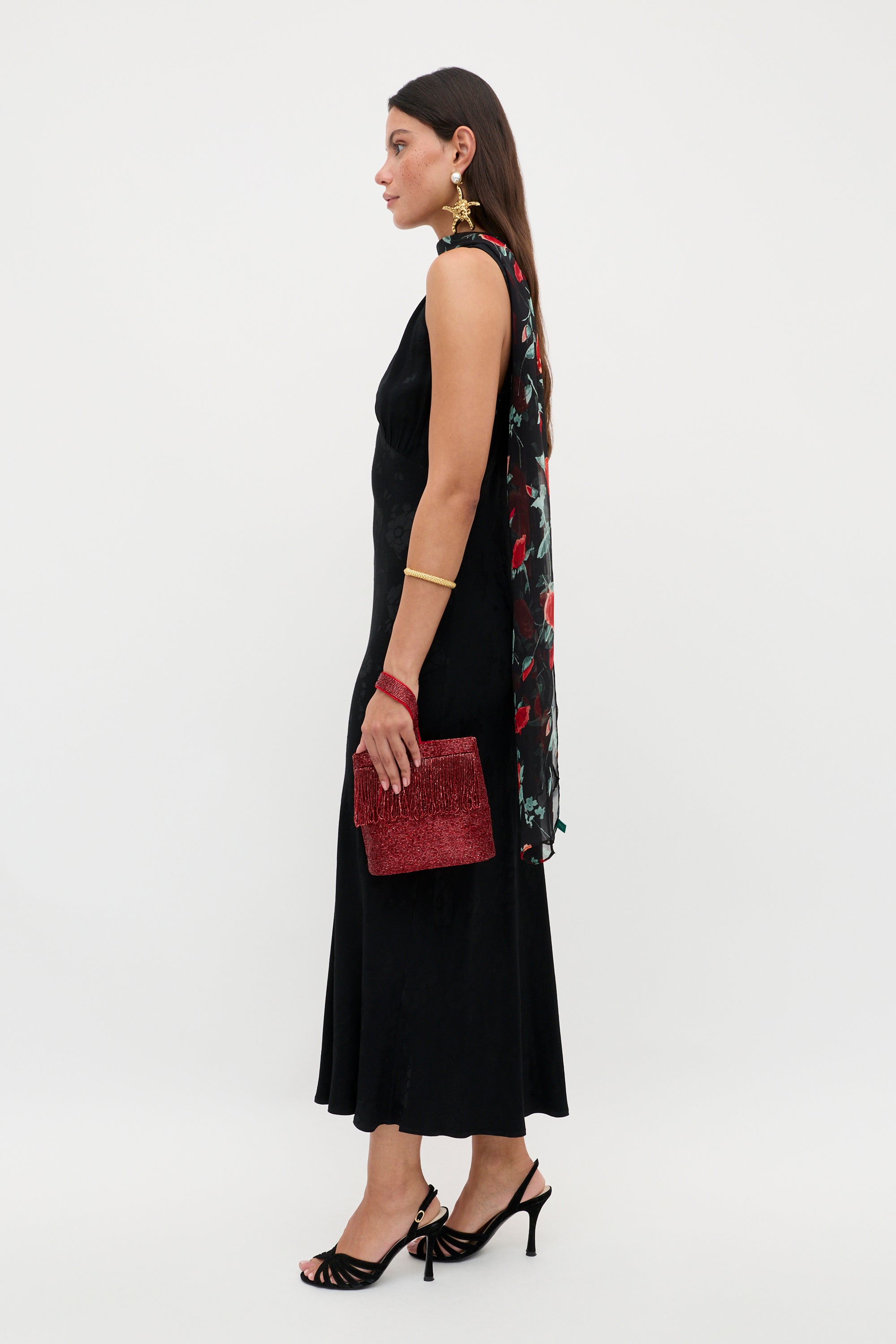 A woman stands in profile wearing the Sandrine - Black Poppy Jacquard sleeveless midi dress, styled with a floral scarf, red textured clutch, black heels, gold earrings, a bracelet, and long dark hair.