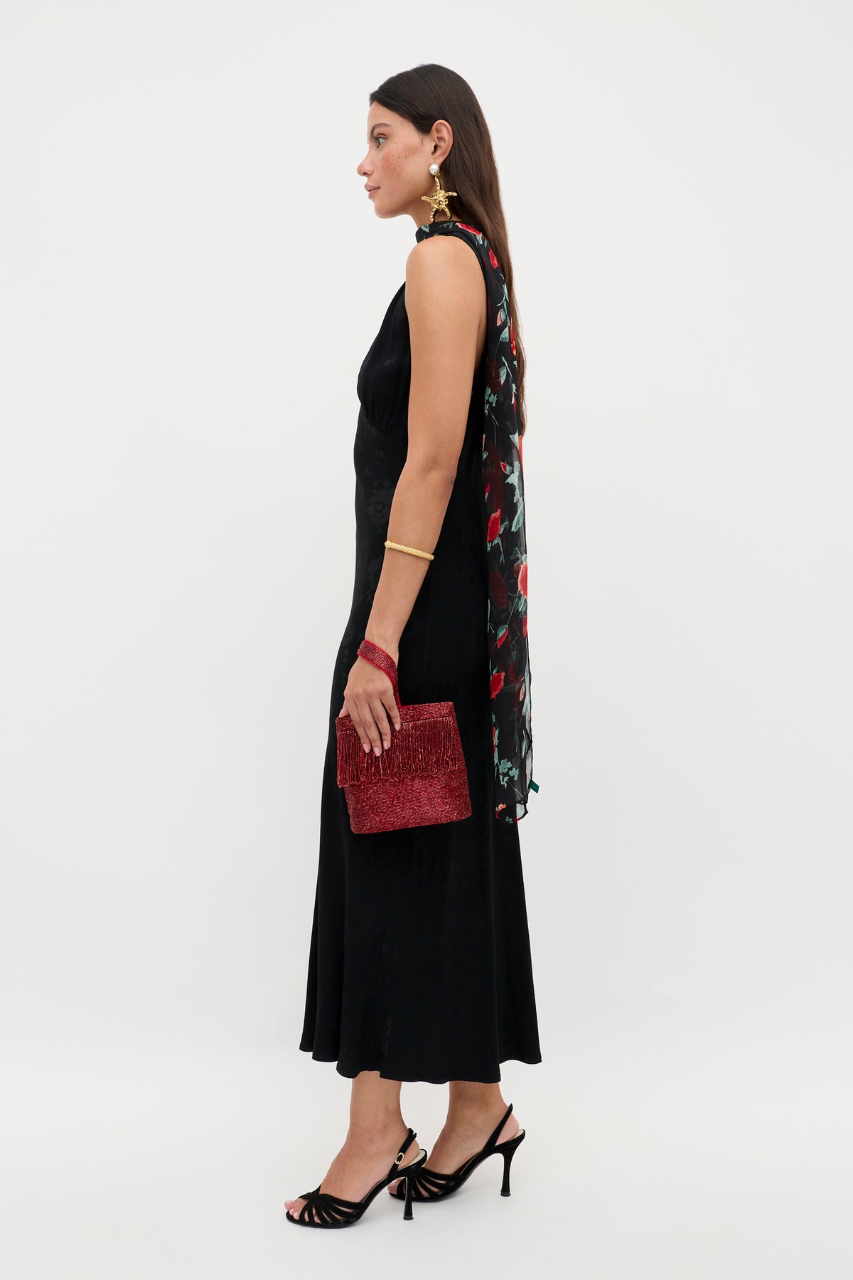 A woman stands in profile wearing the Sandrine - Black Poppy Jacquard sleeveless midi dress, styled with a floral scarf, red textured clutch, black heels, gold earrings, a bracelet, and long dark hair.