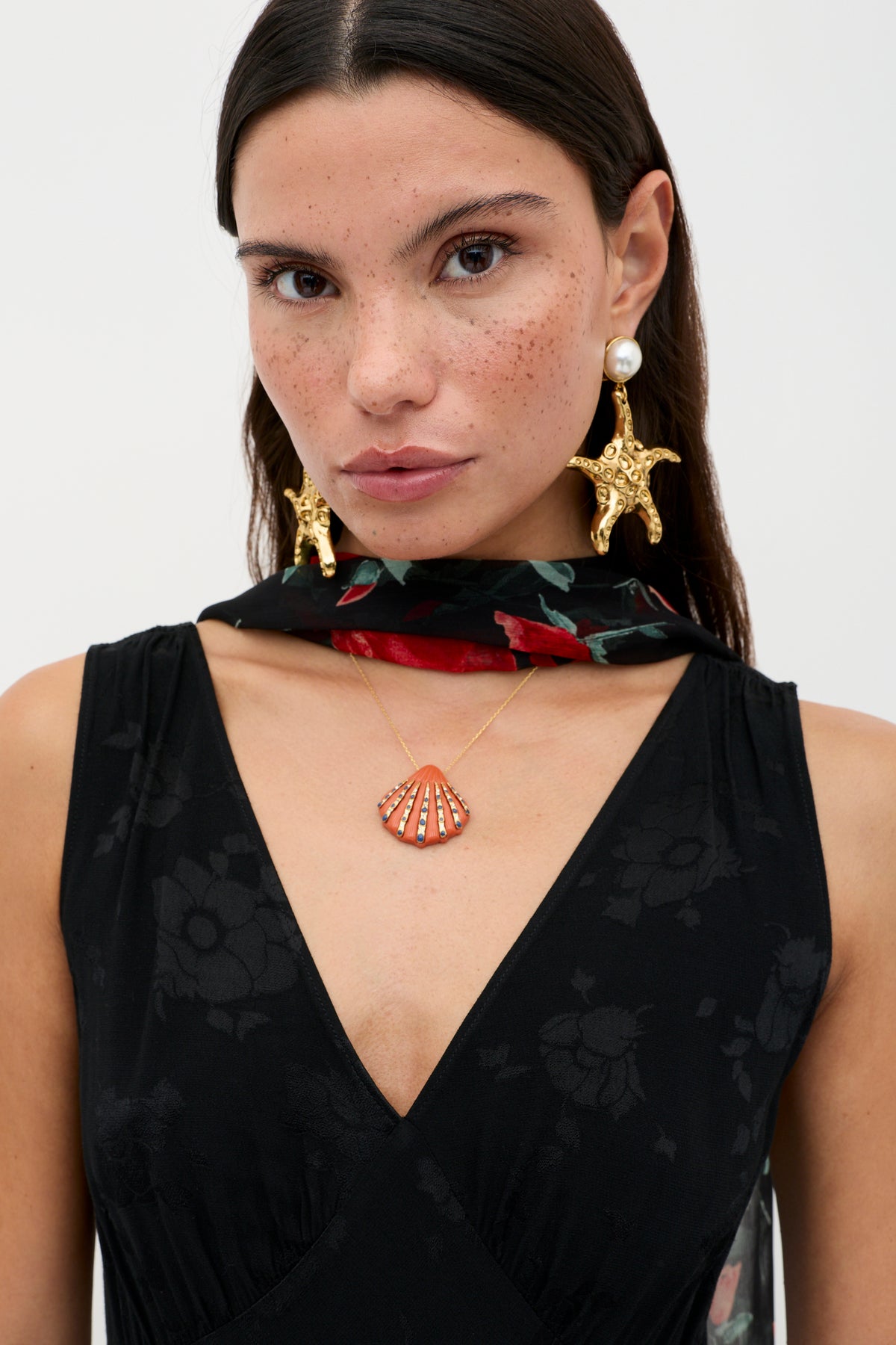 A woman with long dark hair and freckles wears the Sandrine - Black Poppy Jacquard midi dress, a black scarf with red flowers, a gold seashell necklace, and large gold starfish-and-pearl earrings as she looks directly at the camera.