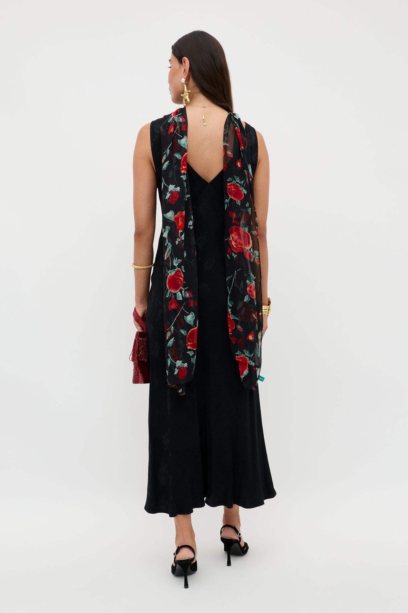 A woman, facing away, wears the Sandrine - Black Poppy Jacquard slip midi dress with a V-back and black heels. She drapes a sheer poppy jacquard scarf over her shoulders, accessorized with gold jewelry and a red purse.