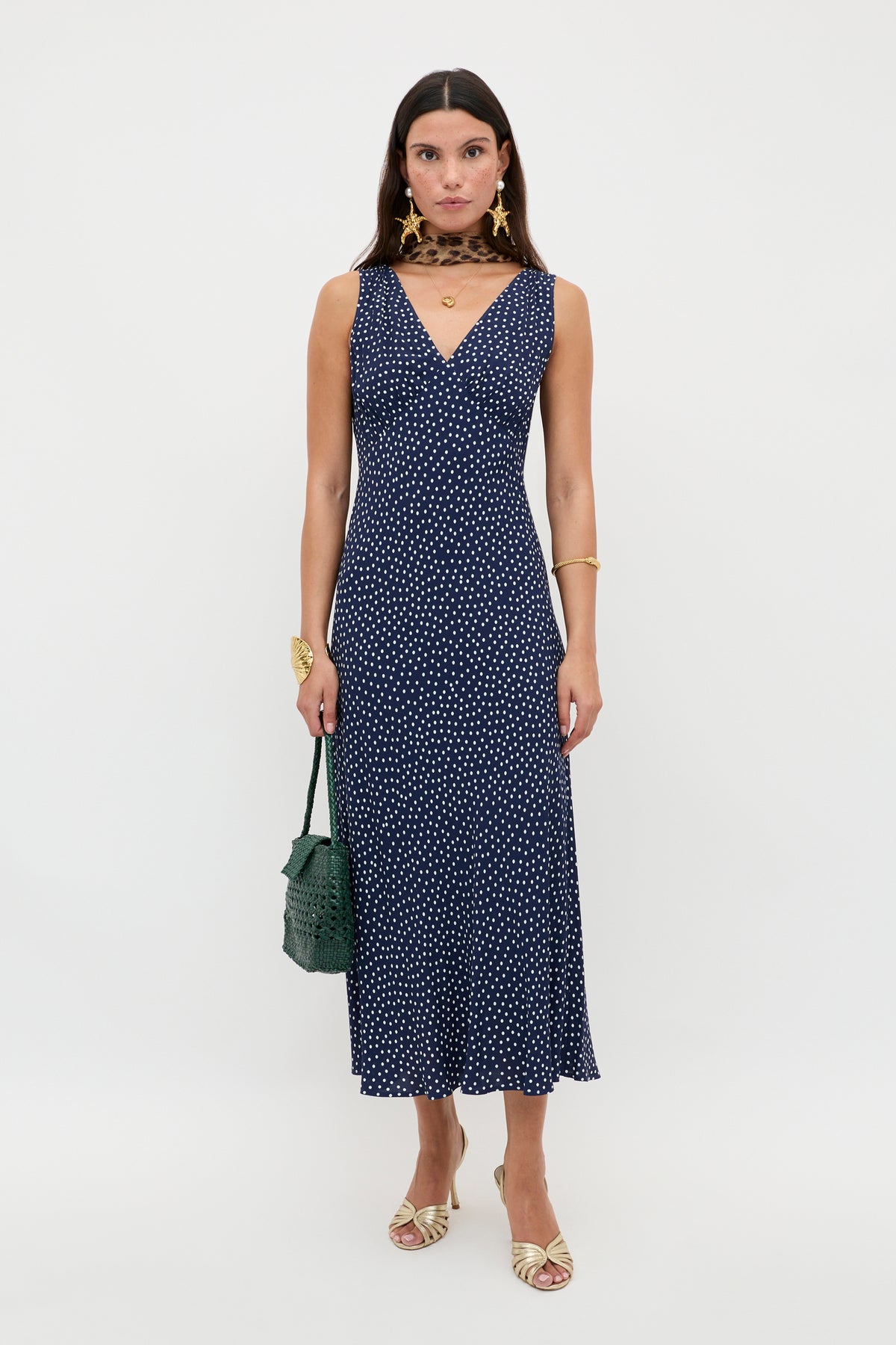 A woman poses against a white background in the Sandrine - Mini Dot Navy dress, featuring a vintage-inspired navy polka-dot print and V-neckline. She accessorizes with gold jewelry, a green handbag, and cream strappy heeled sandals.