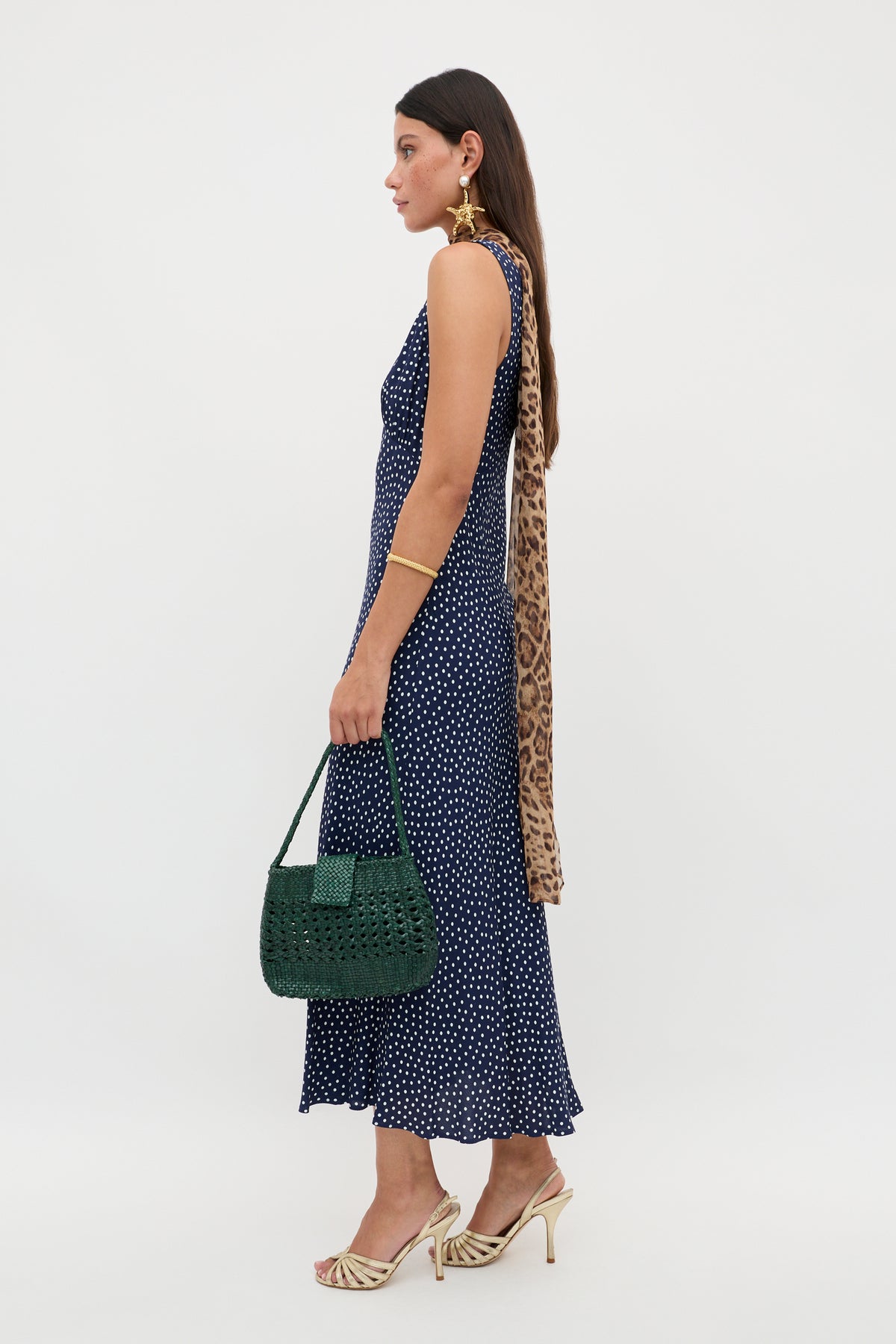 A woman in the Sandrine - Mini Dot Navy dress pairs it with beige heeled sandals, gold jewelry, a green woven handbag, and a leopard-print scarf in her long hair, standing against a white background.