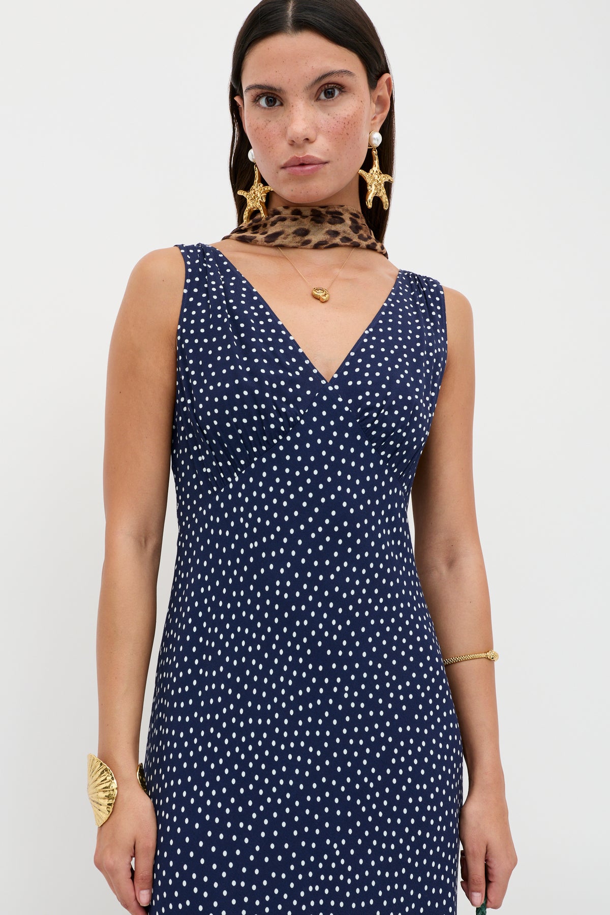 Wearing the Sandrine - Mini Dot Navy vintage-inspired bias-cut slip dress with white polka dots, gold jewelry, and a brown leopard-print scarf, a person poses against a plain white background.