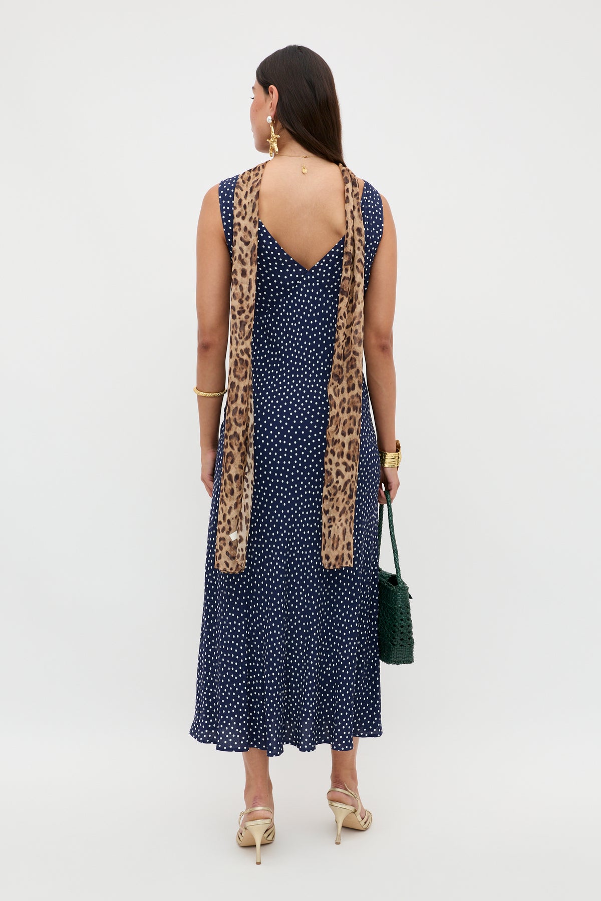 A woman faces away, wearing the Sandrine - Mini Dot Navy vintage-inspired, bias-cut slip dress with a polka-dot print, beige heels, a leopard print scarf over her shoulders, a green woven bag, and gold jewelry.