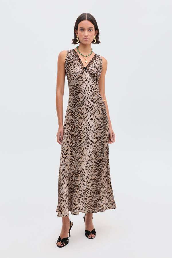 A woman wearing the sleeveless, v-neck Sandrine - Antelope Spot Black slip dress by RIXO, paired with black high-heeled sandals and a green beaded necklace, stands against a plain white background.