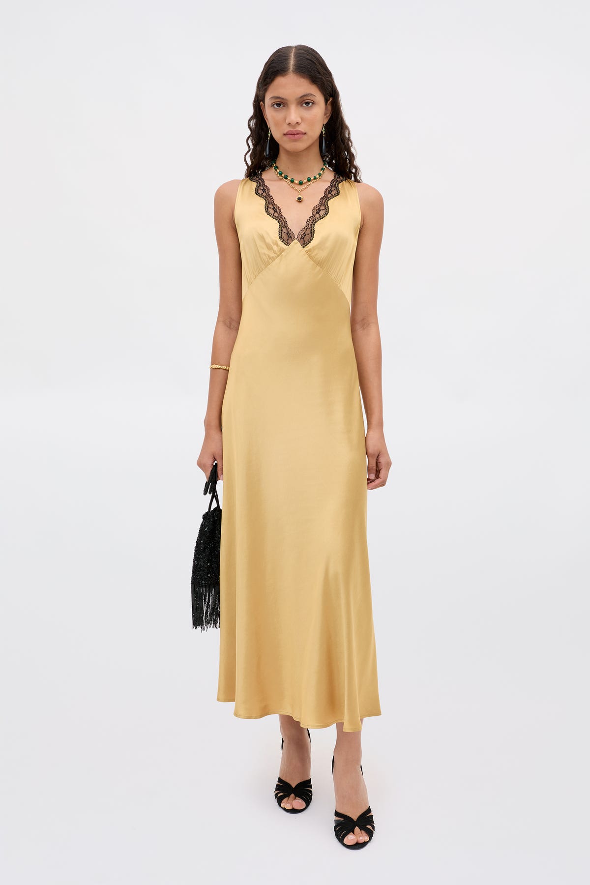 A woman in the Sandrine - Gold long, sleeveless yellow satin dress with black lace trim, paired with black high-heeled sandals, a fringed black handbag, and layered necklaces stands against a plain white background.