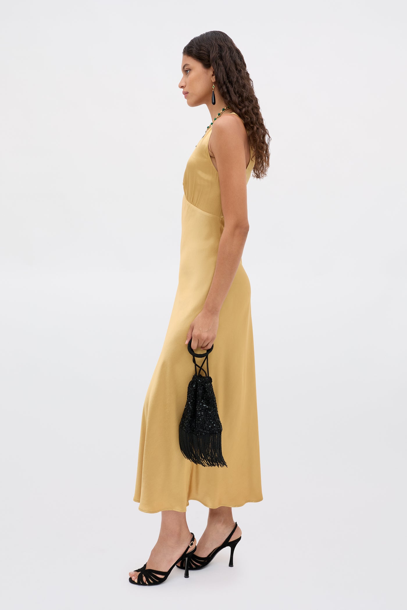 A woman with long curly hair wears the Sandrine - Gold sleeveless midi dress and black heeled sandals, holding a small black fringed handbag, standing in profile against a plain light background.