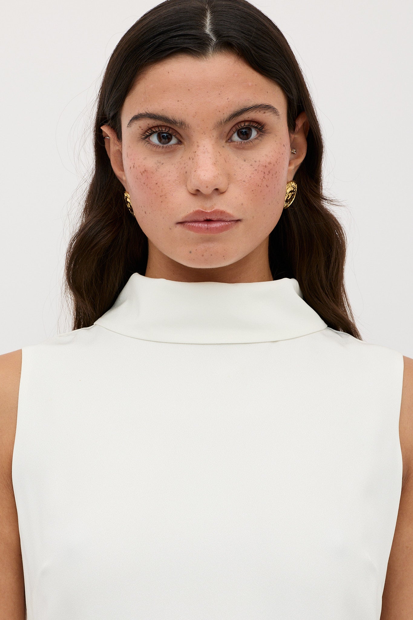 A woman with long brown hair and freckles models the Candice - Satin Back Silk Crepe: a sleeveless, high-neck white top with a subtle sixties-inspired shift dress silhouette, against a plain light background.