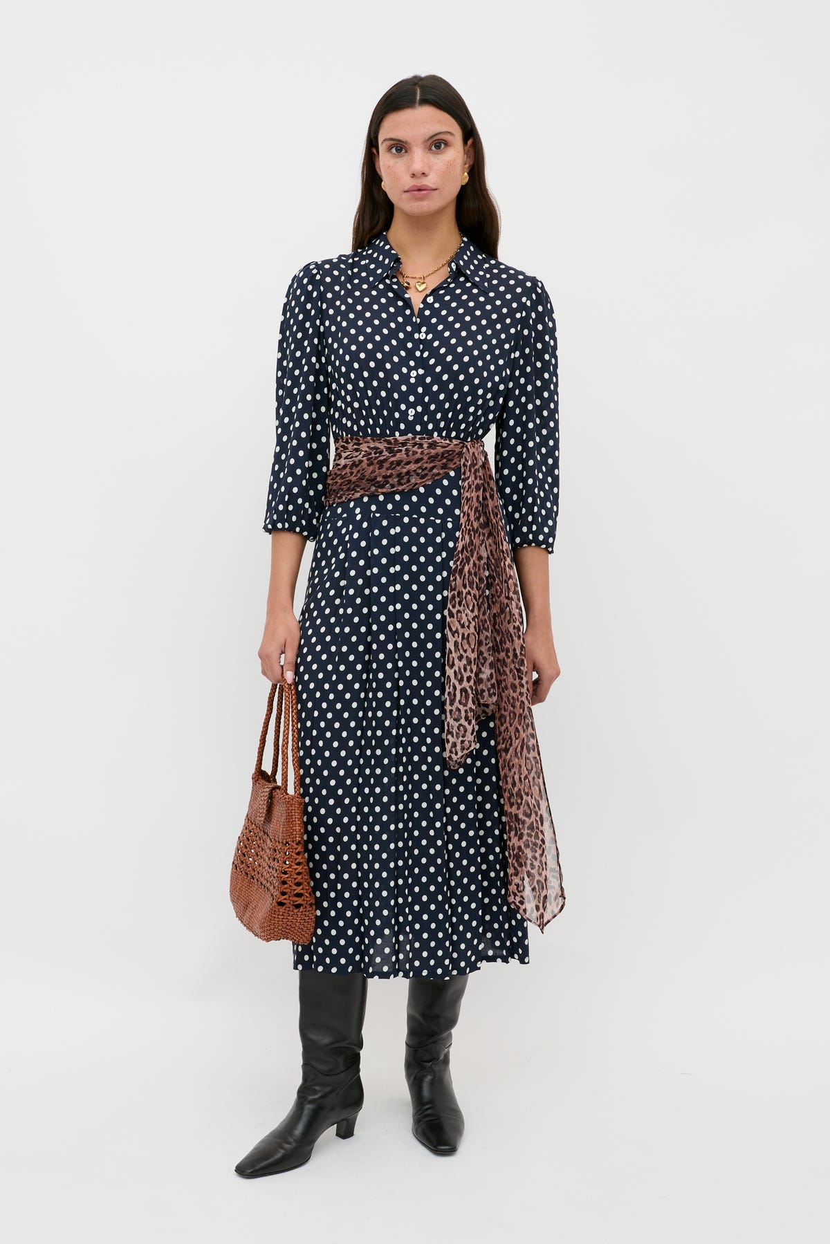 A woman stands against a white background wearing the Izzy - Runway Spot Navy dress, styled with a leopard print sash, black knee-high boots, and holding a brown woven handbag.